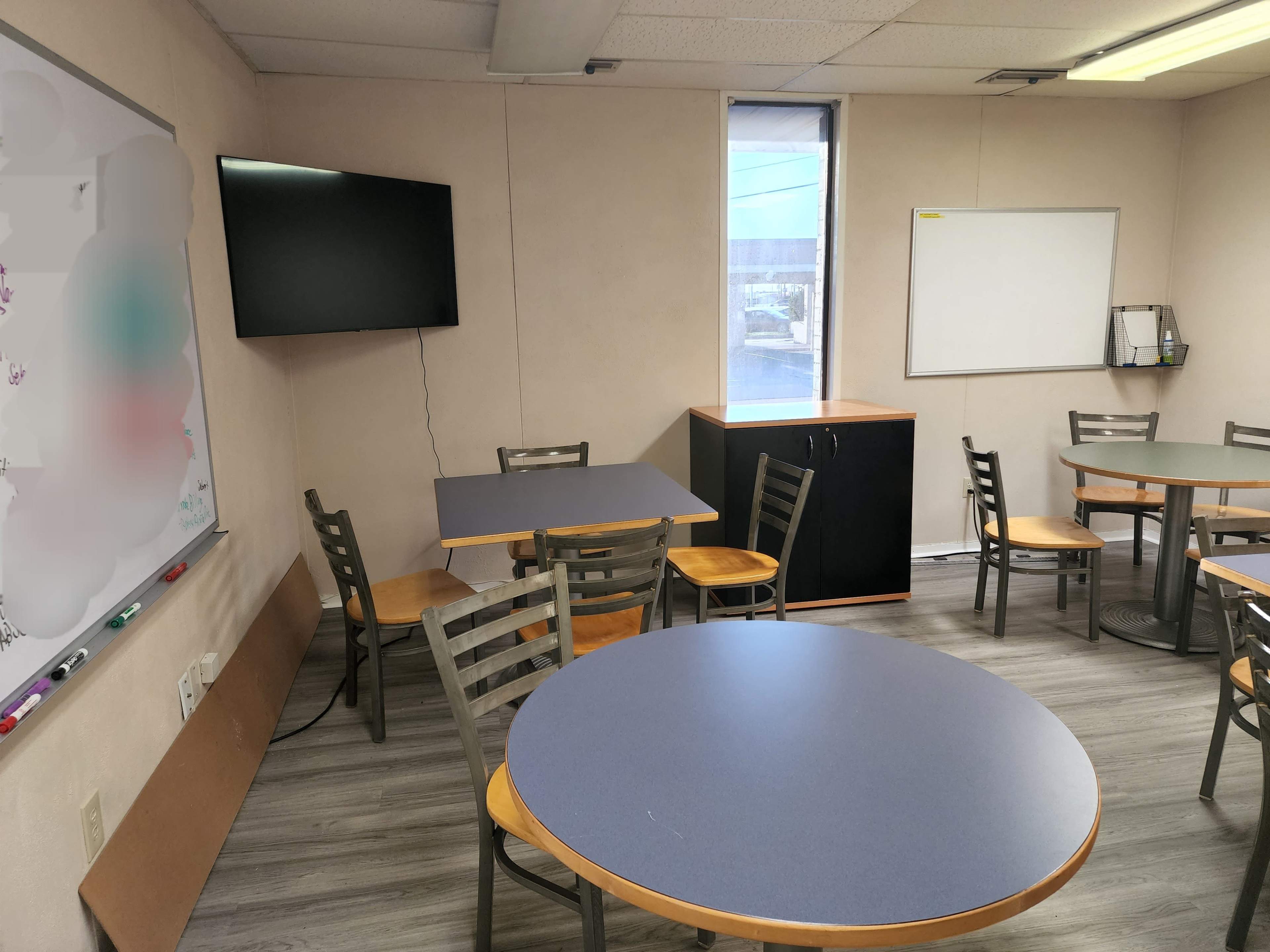 A classroom with multiple round tables and chairs, a whiteboard, a television mounted on the wall, and a black cabinet in the corner.