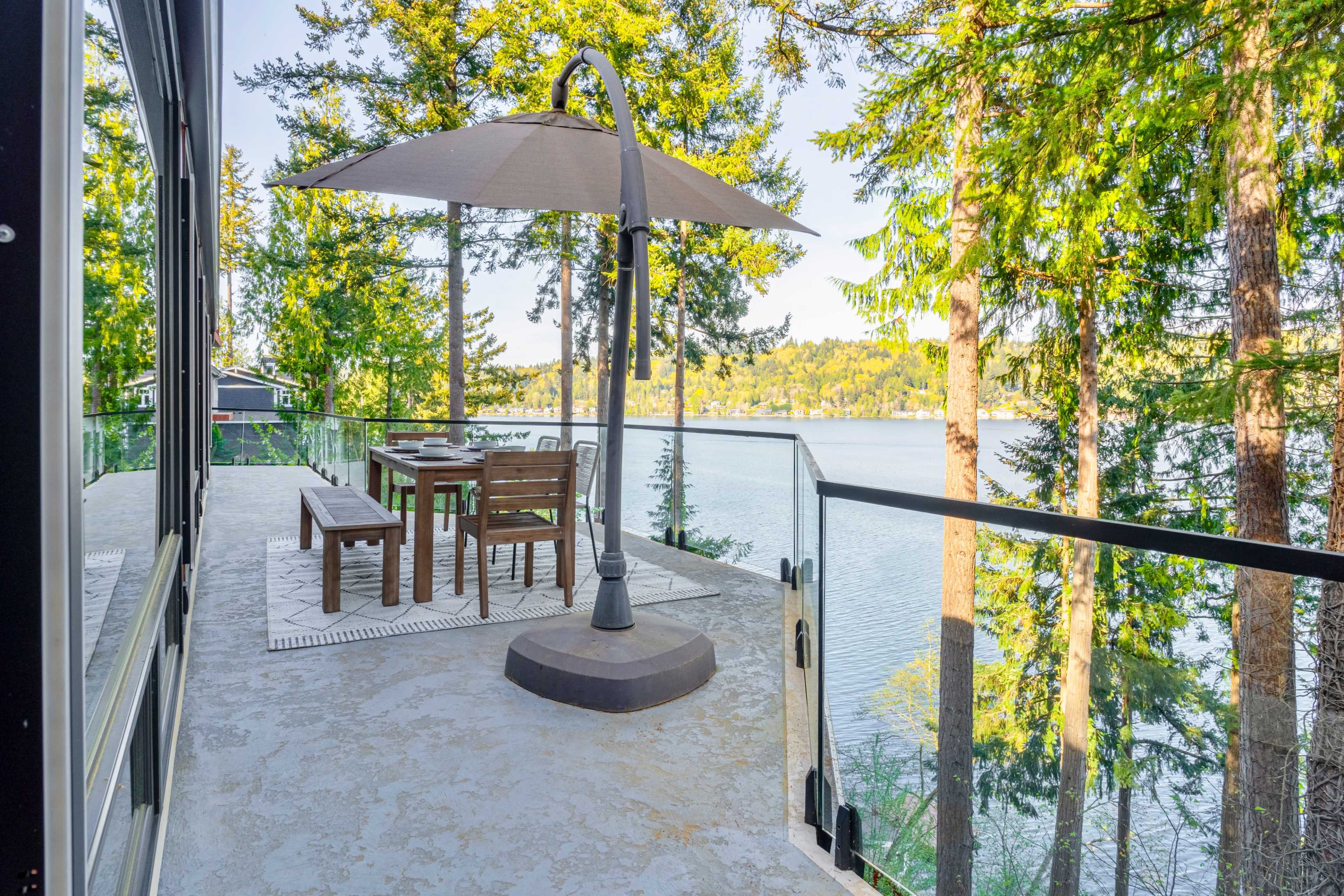 A patio with a table and chairs is set near a lake, featuring a large umbrella and surrounded by trees.