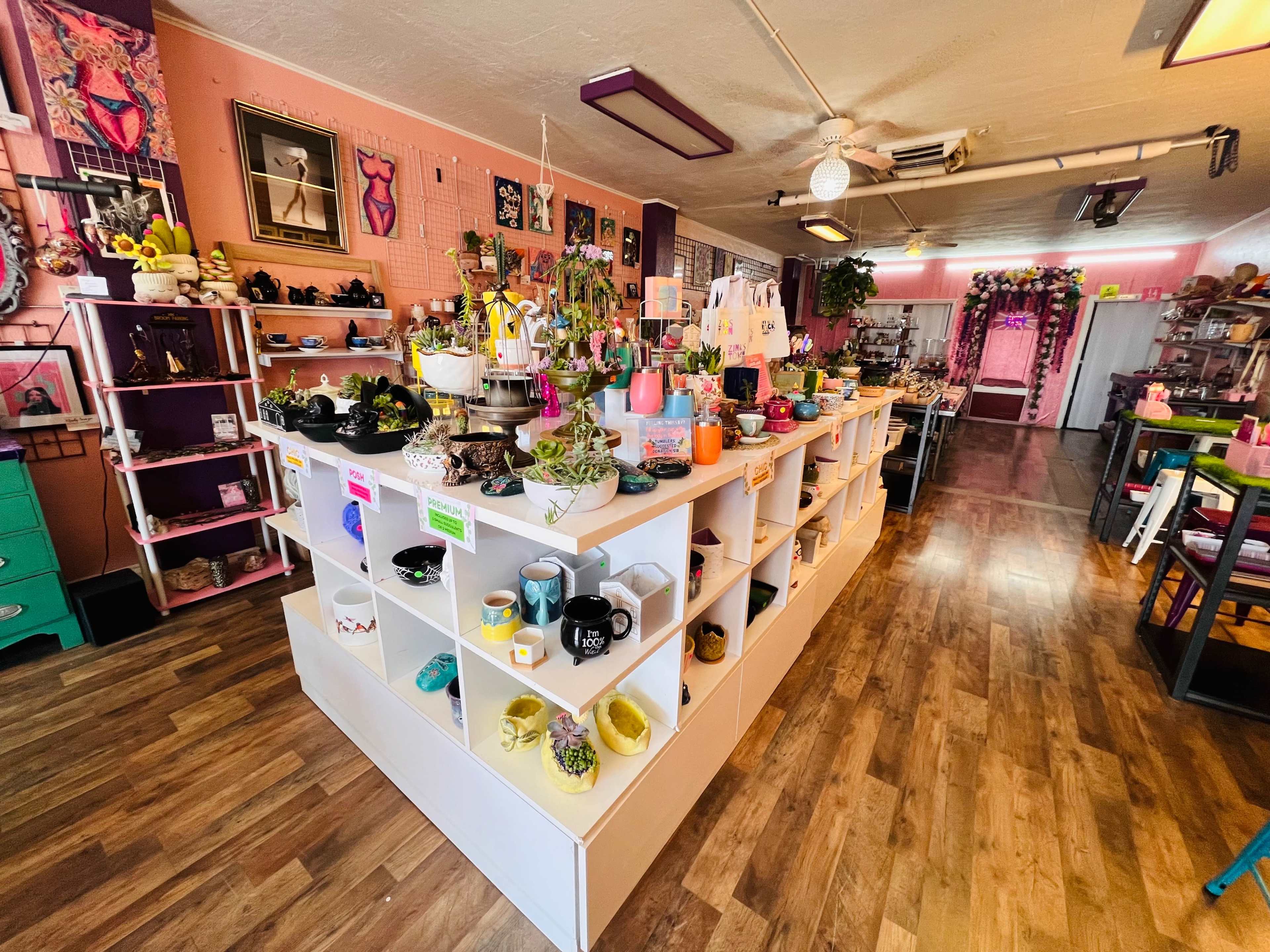 A vibrant retail space featuring a white display counter filled with various decorative items, plants, and colorful merchandise against a backdrop of pink walls and eclectic decor.