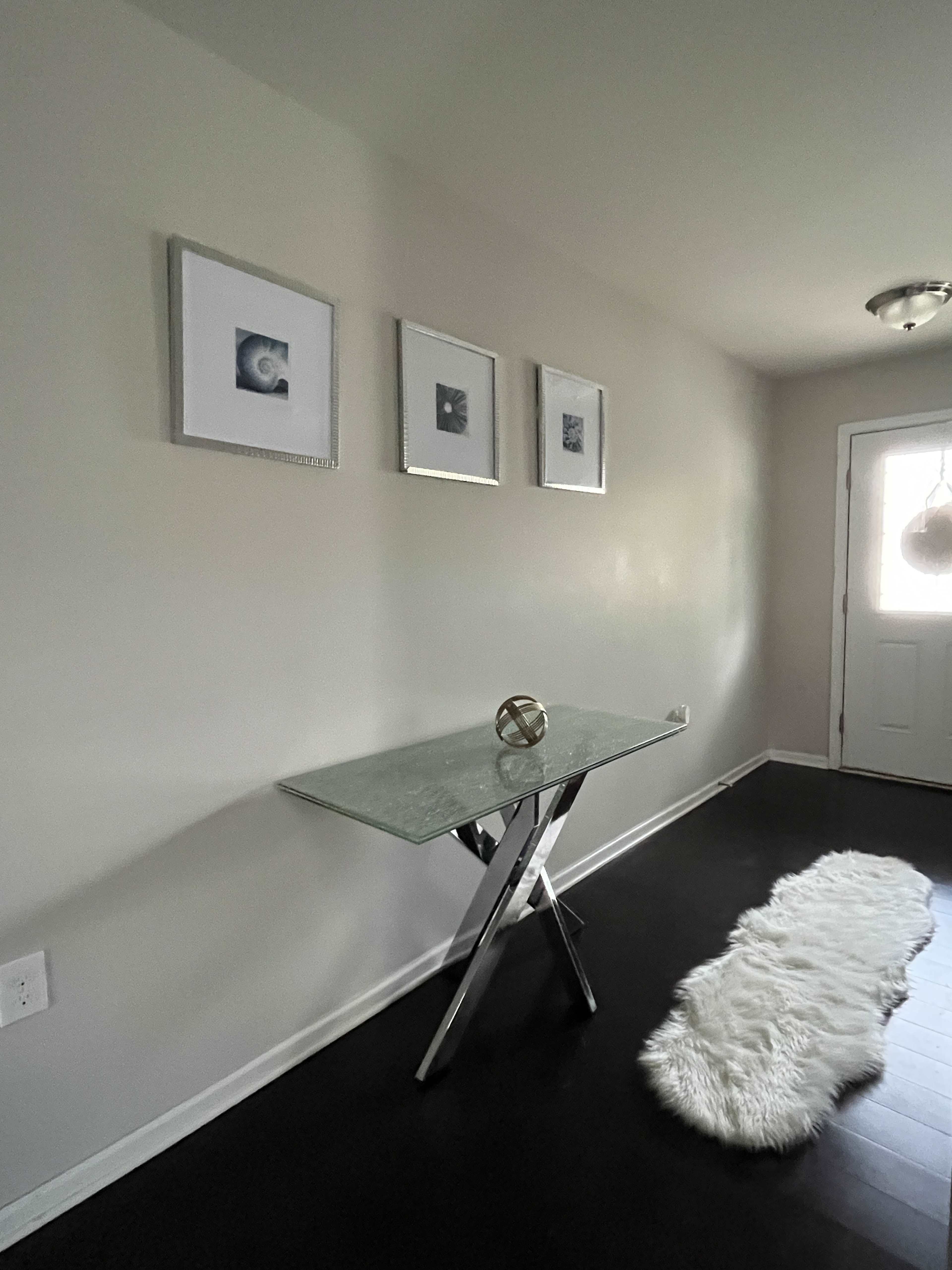A glass-top table with a metallic base sits against a light-colored wall, accompanied by three framed pictures and a white faux-fur runner leading to a door.