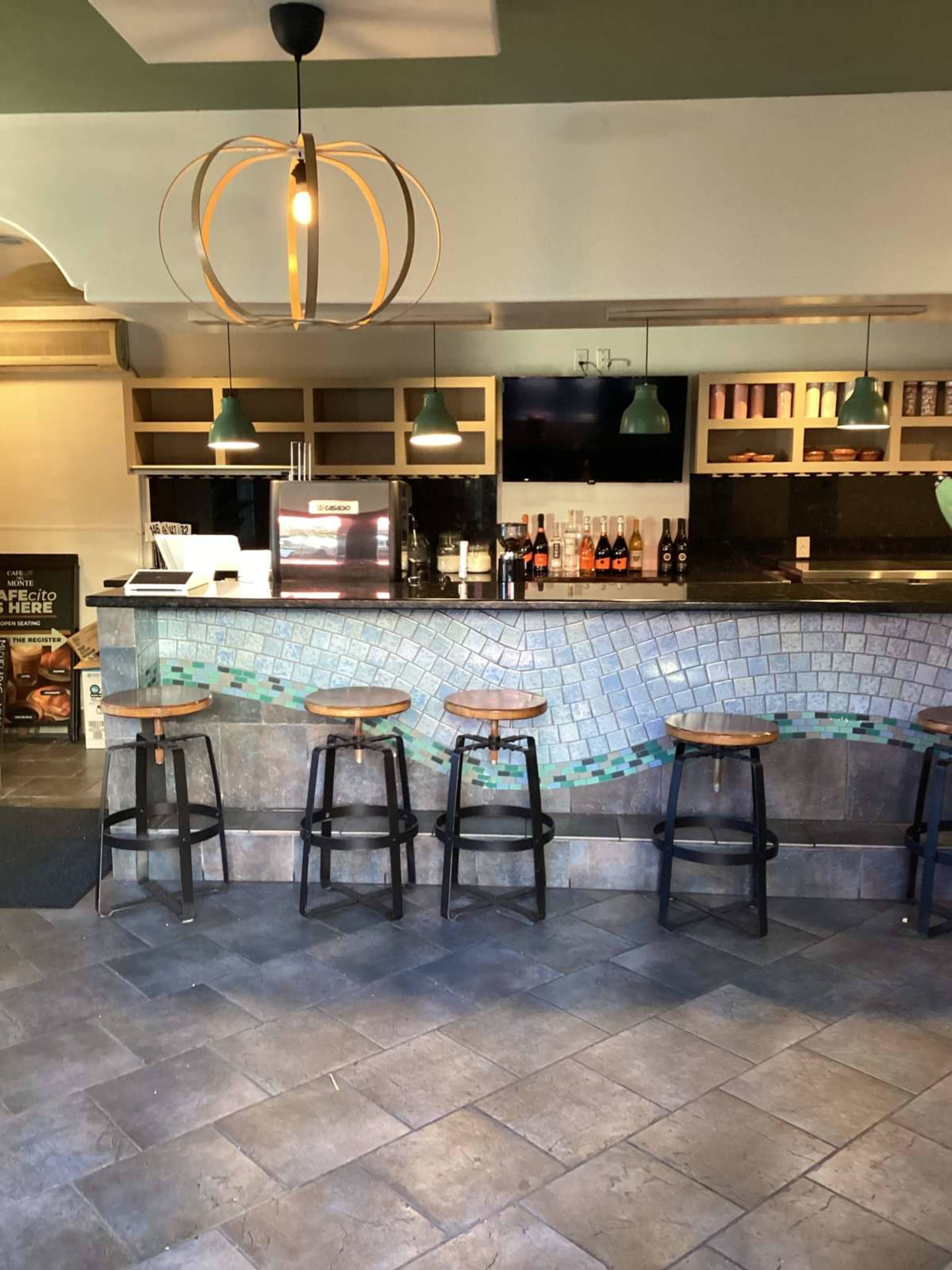 The image shows a bar area featuring a counter with wooden stools, a backlit display of bottles, and pendant lighting above a tiled surface.