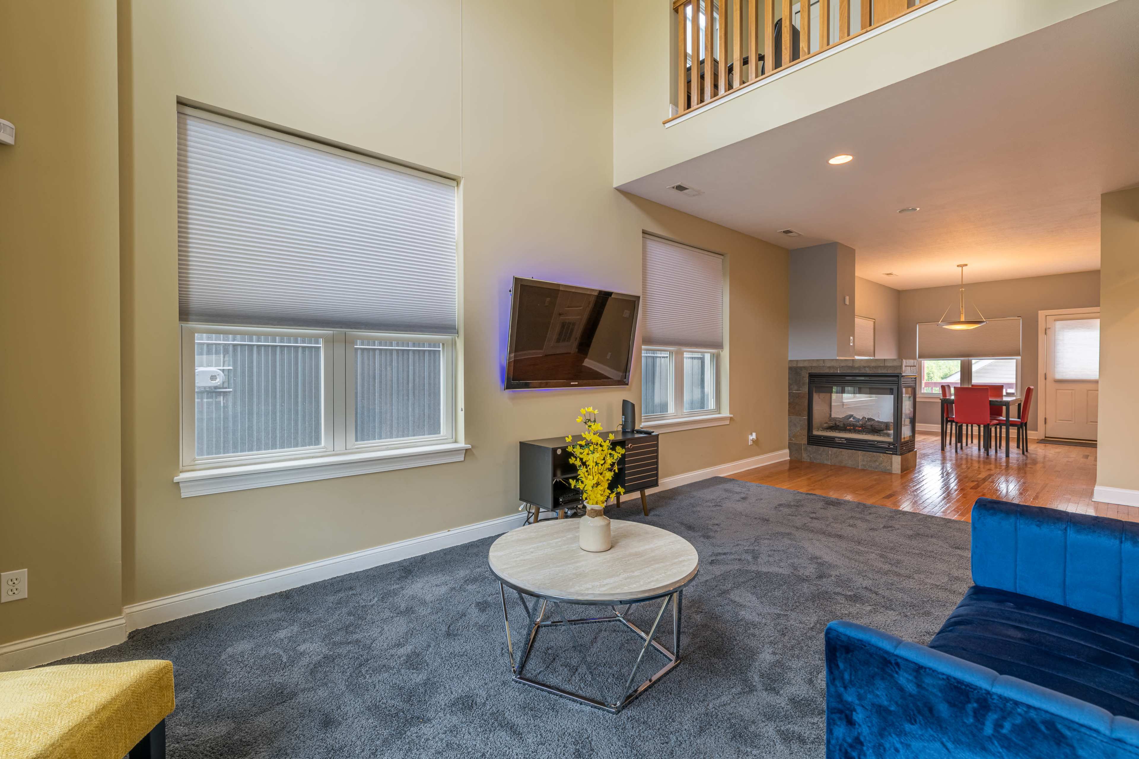 A living room features a beige wall, blue furniture, a round coffee table, and a flat-screen TV mounted on the wall, with large windows allowing natural light.