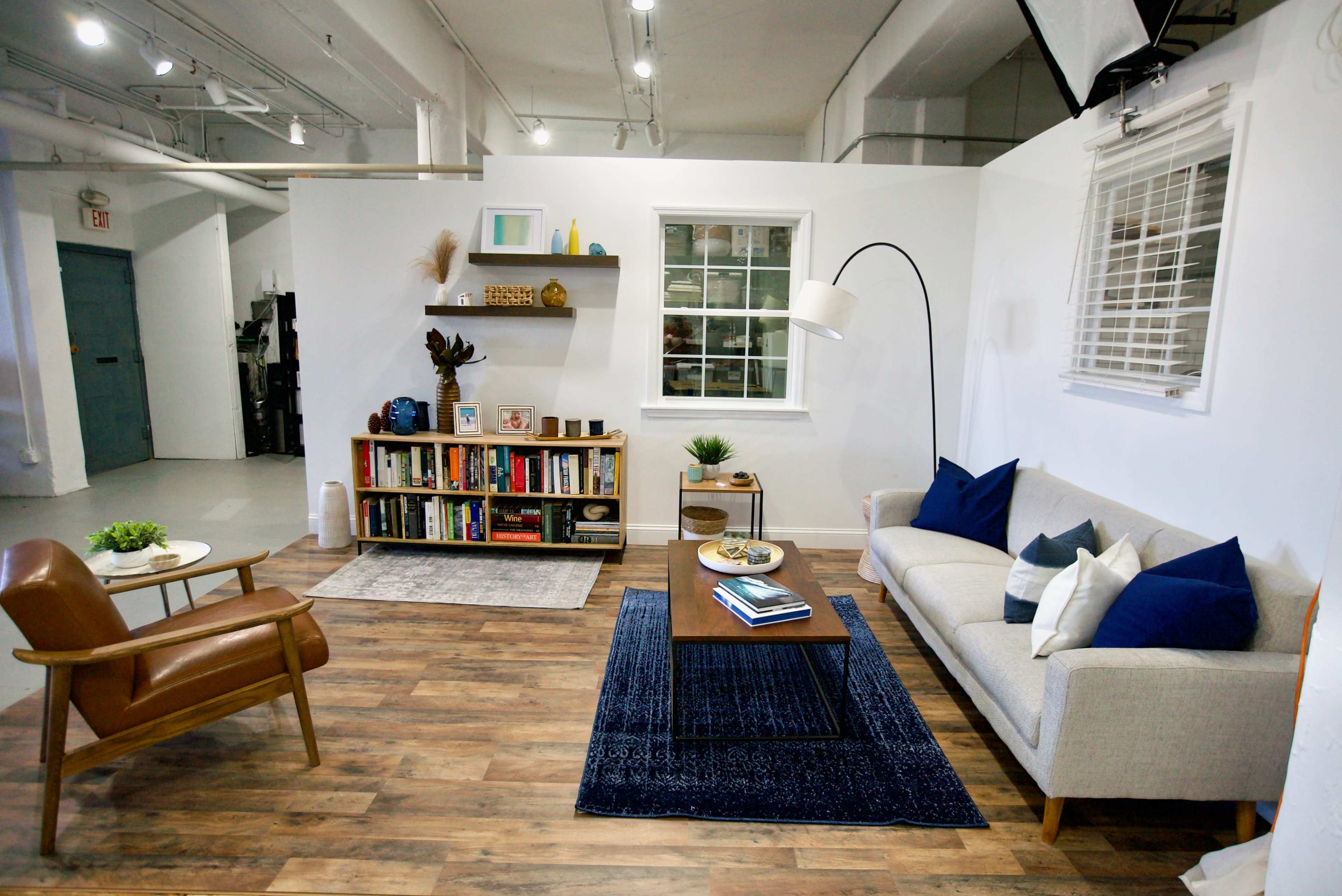 The image shows a modern living room setup with a beige sofa, a wooden coffee table on a blue rug, a bookshelf filled with books, and a mid-century modern chair.