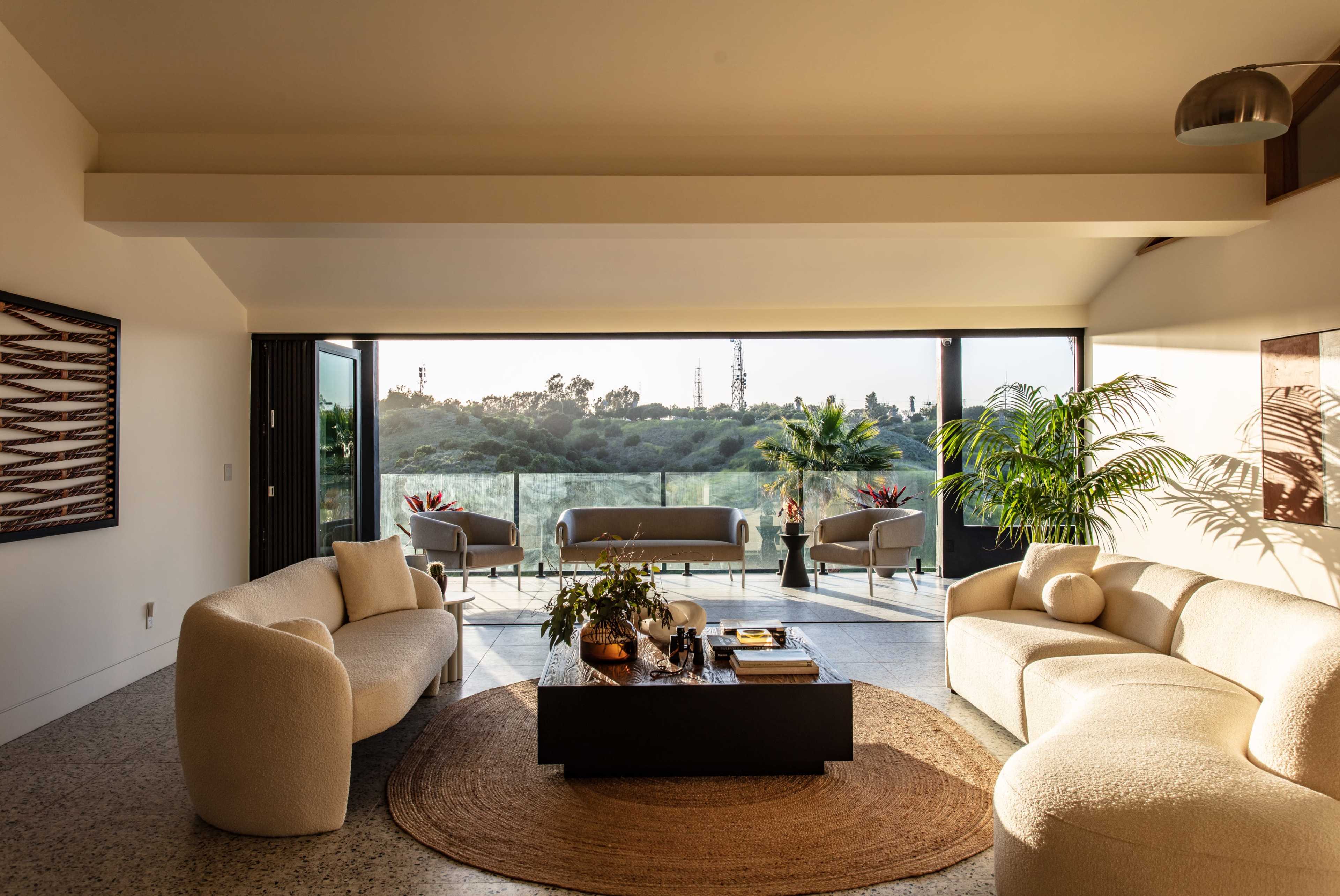 A modern living room features curved sofas and a coffee table overlooking a scenic balcony with greenery in the background.