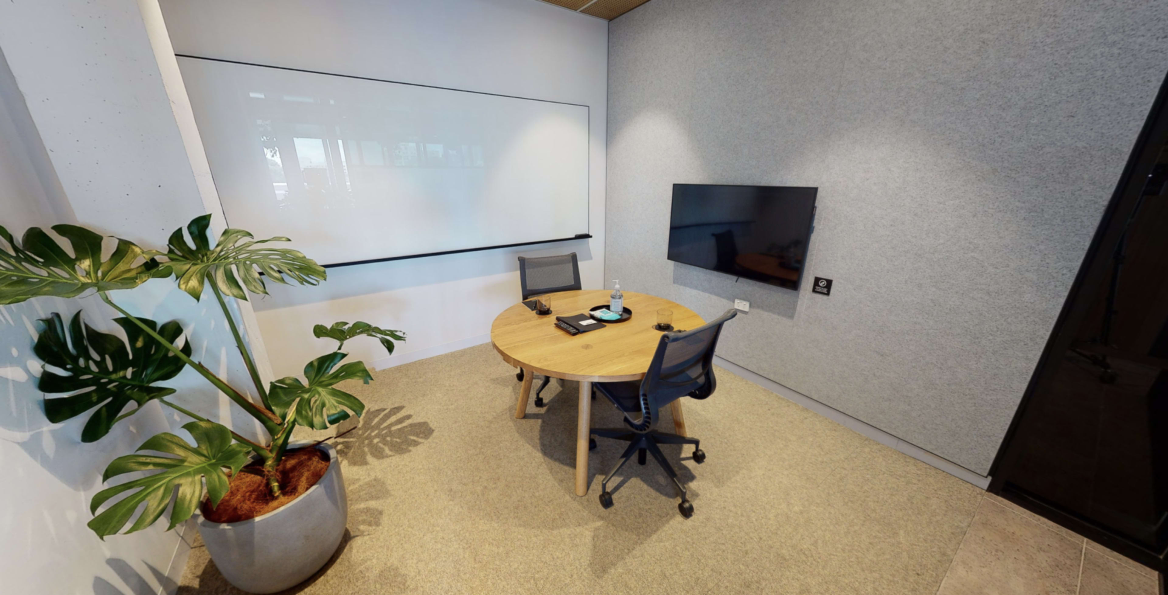 A minimalistic meeting room features a round wooden table with two chairs and a large wall-mounted screen, accompanied by a potted plant in the corner.