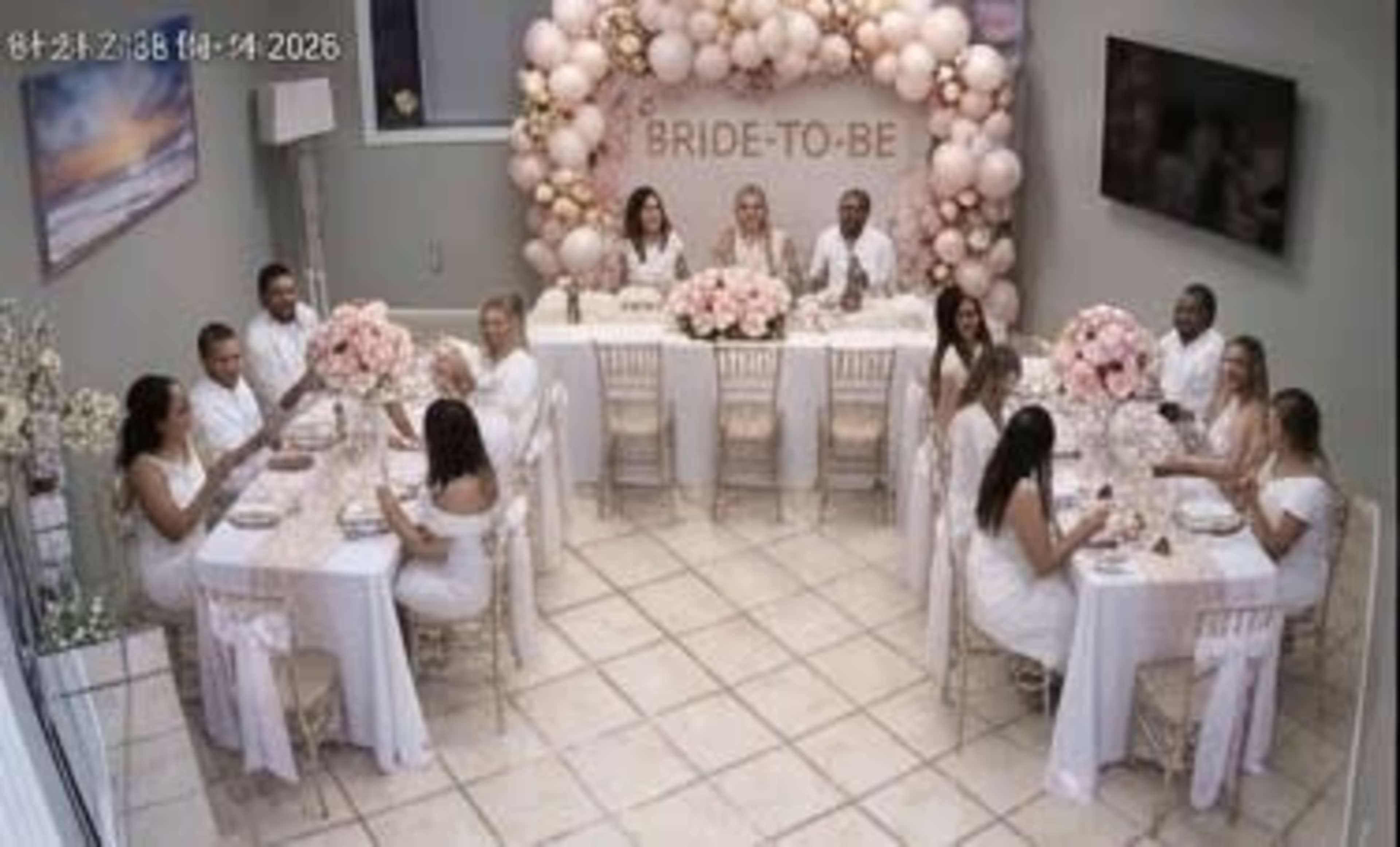 A group of people wearing white attire are gathered at a decorated table with floral arrangements for a celebration labeled "BRIDE-TO-BE."