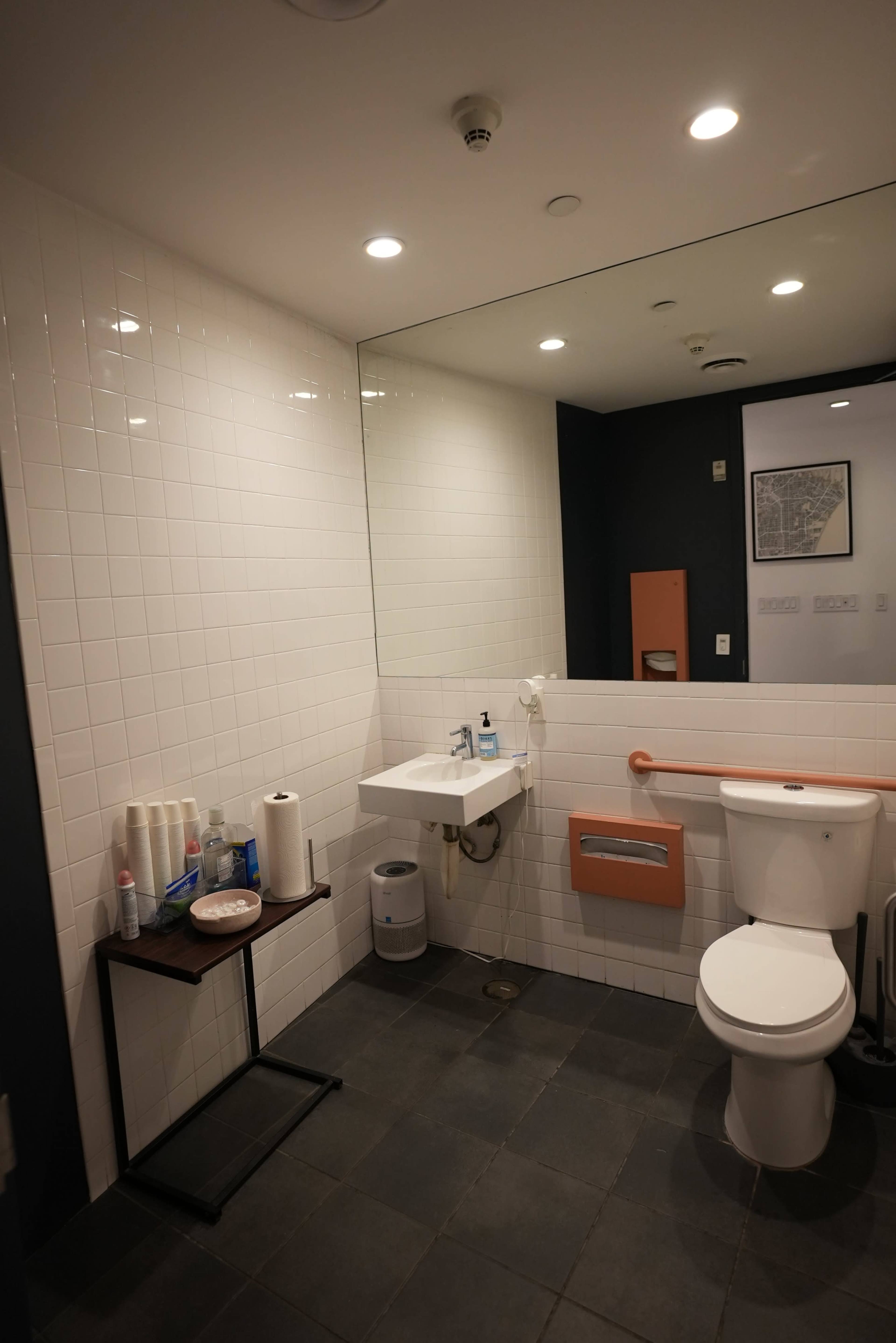 The image shows a clean bathroom with a sink, a toilet, and a large mirror, featuring tiled walls and a dark floor.
