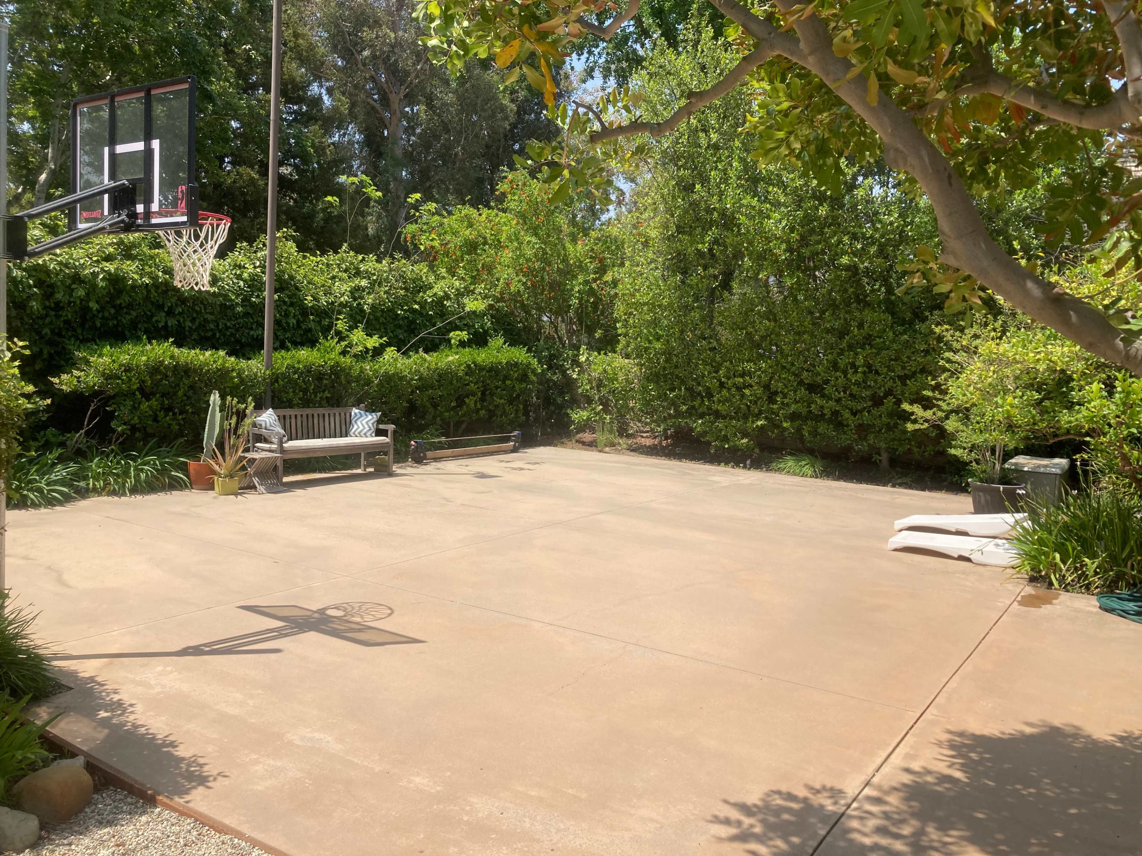 The image shows a backyard basketball court surrounded by lush greenery, with a bench and some equipment on one side.
