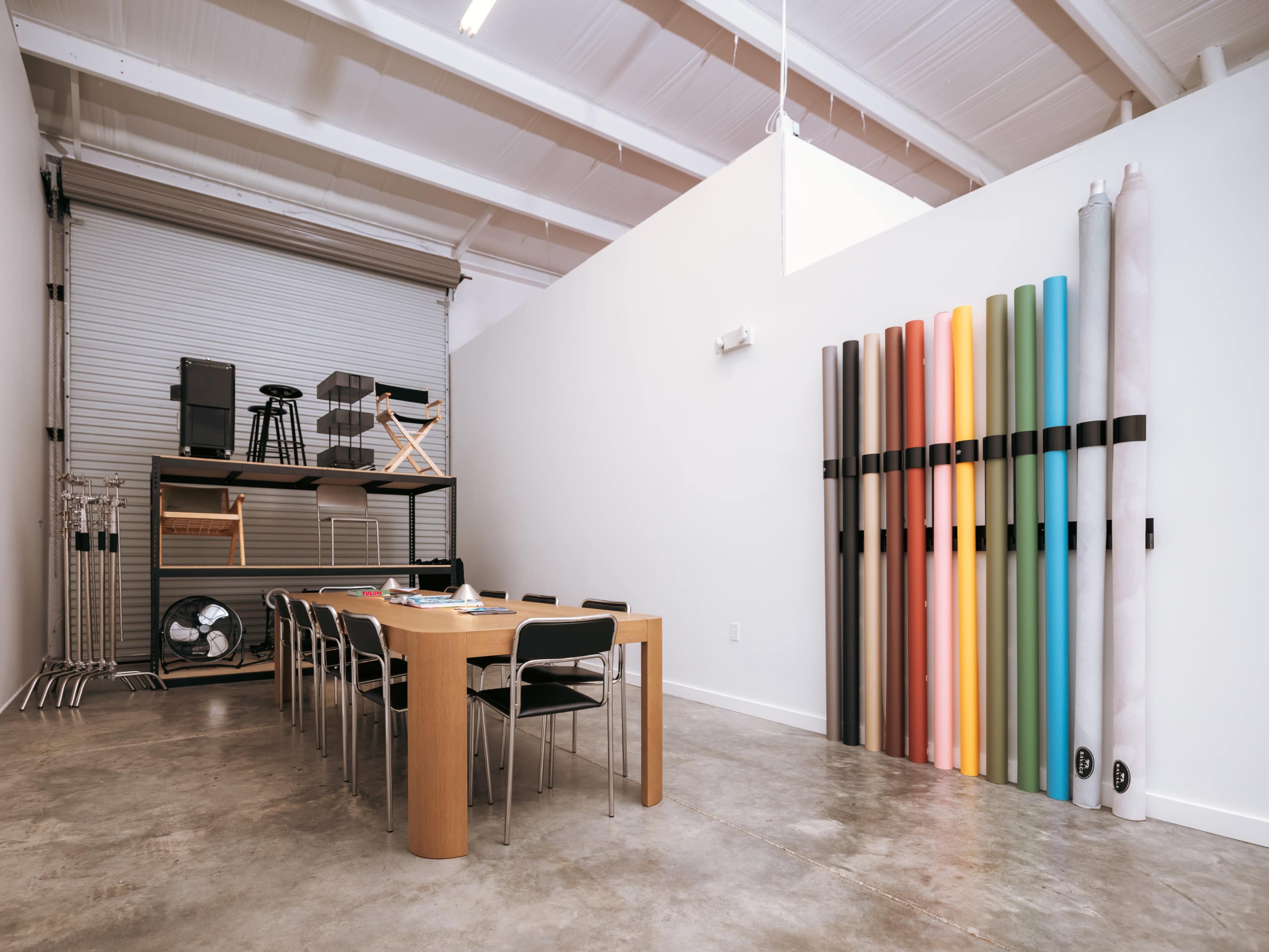 A spacious room features a large wooden table surrounded by black chairs, with rolled-up colorful materials displayed on the wall and storage shelving behind.