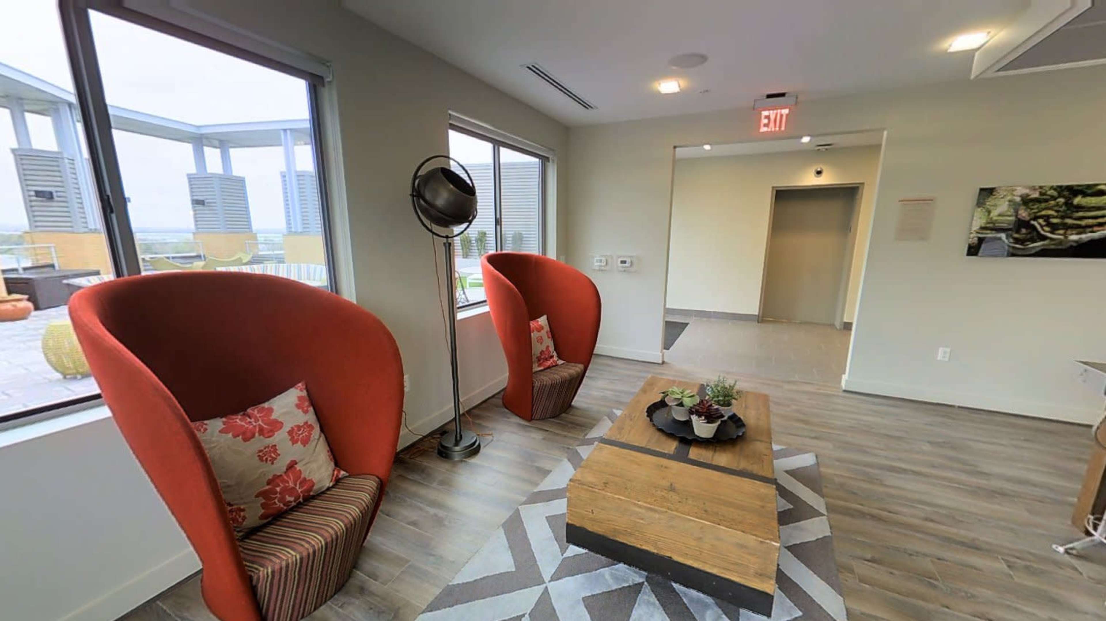 The image shows a modern lounge area featuring two red, upholstered chairs, a wooden coffee table, and large windows allowing light into the space.