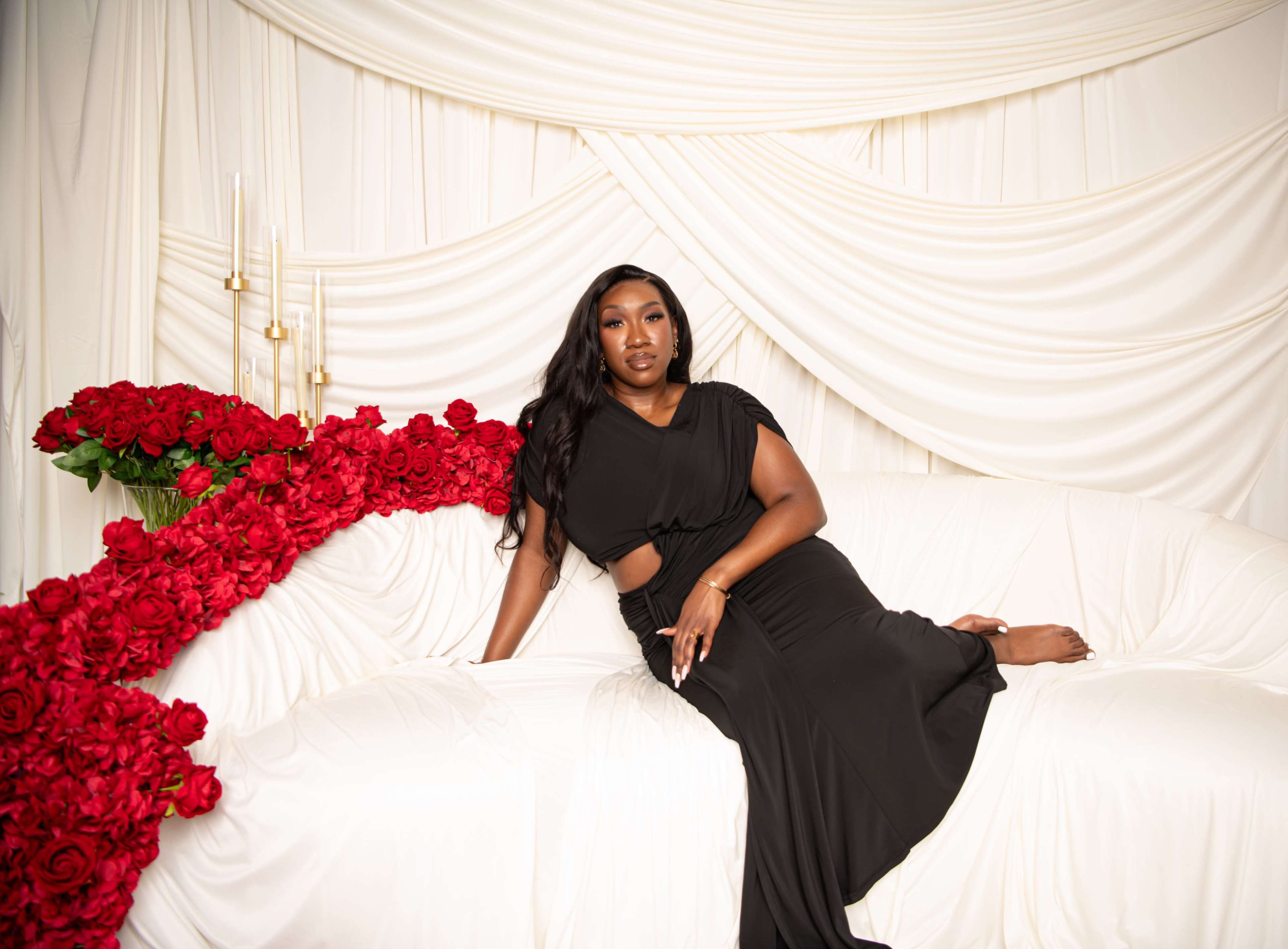 A woman in a black dress sits on a white couch surrounded by a cascade of red roses against a draped white backdrop.