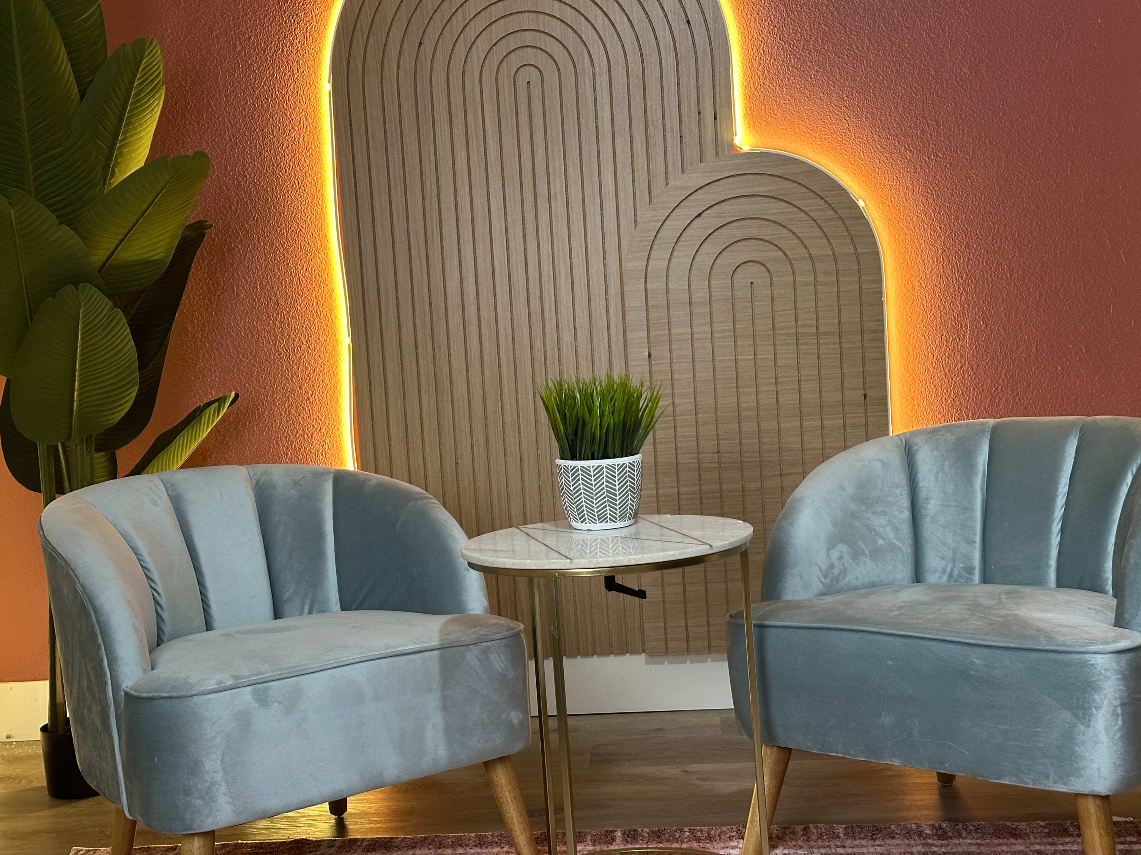 The image features two light blue velvet chairs next to a small round table, with a potted plant and a wooden wall design illuminated by soft lighting in the background.