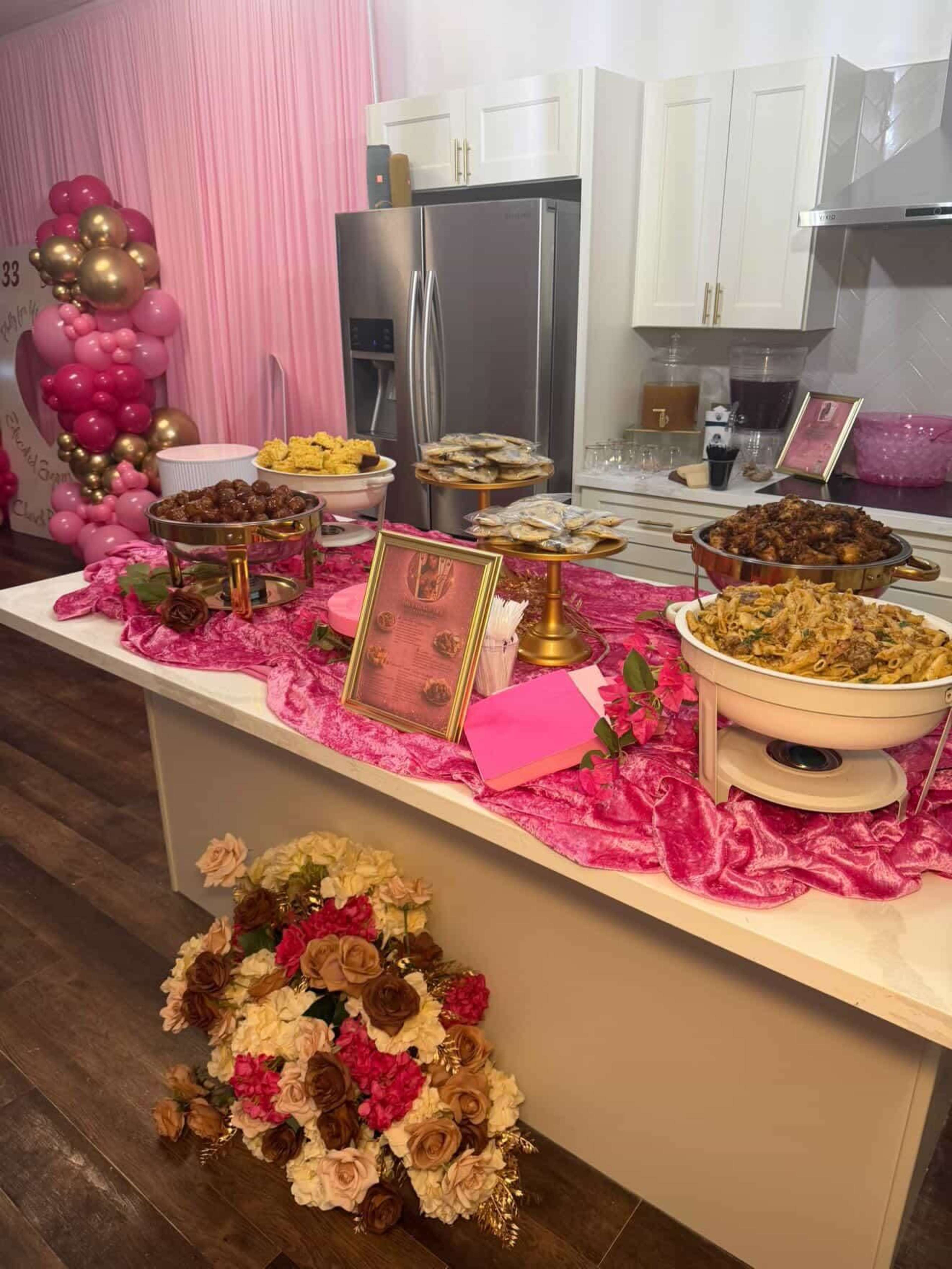 The image shows a decorated table with various food dishes, framed signs, and floral arrangements, set against a backdrop of pink balloons in a modern kitchen.