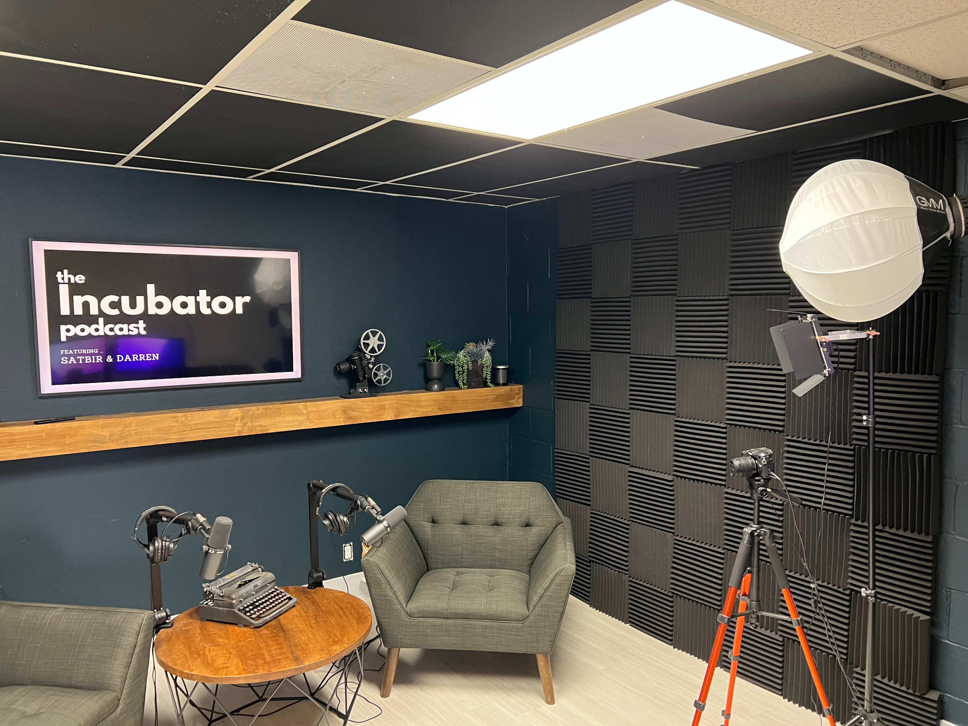 A podcast studio featuring a dark blue wall, two gray chairs, a wooden table, a vintage typewriter, and lighting equipment, with the title "the Incubator podcast" displayed on a screen.