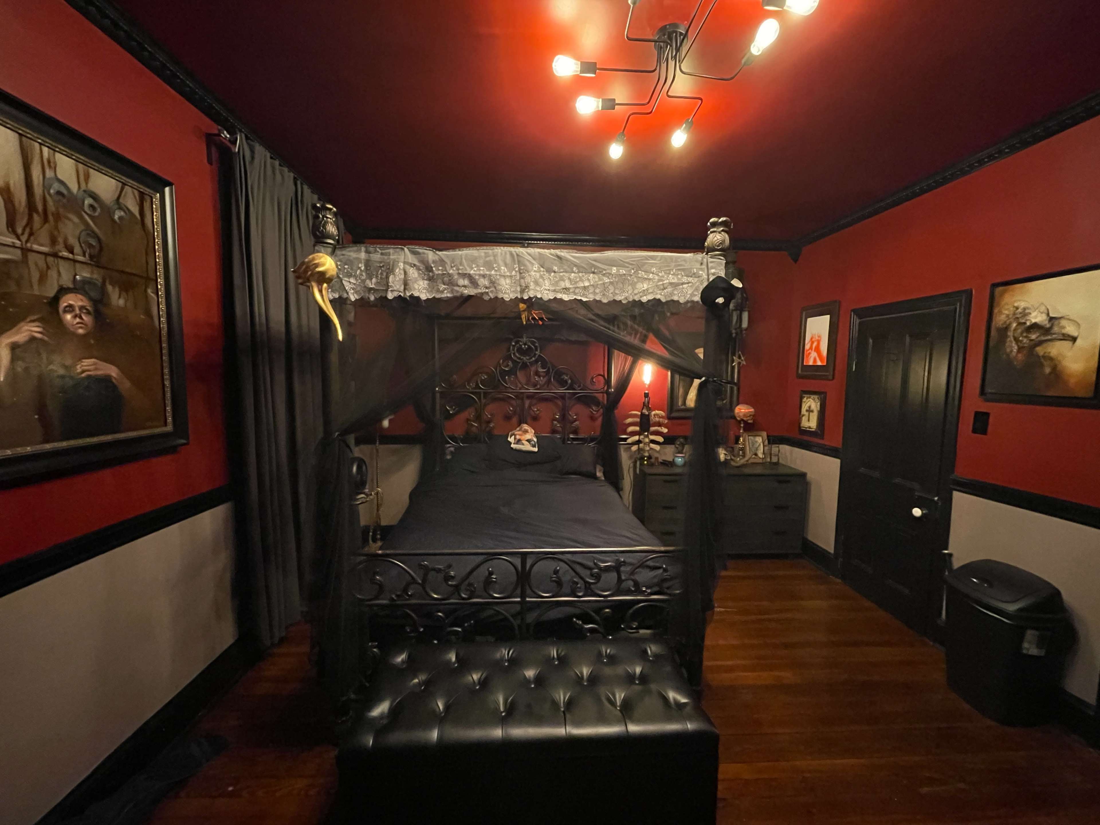 A dimly lit bedroom with a canopy bed, dark walls adorned with framed artwork, and a dresser beside a window with draped curtains.