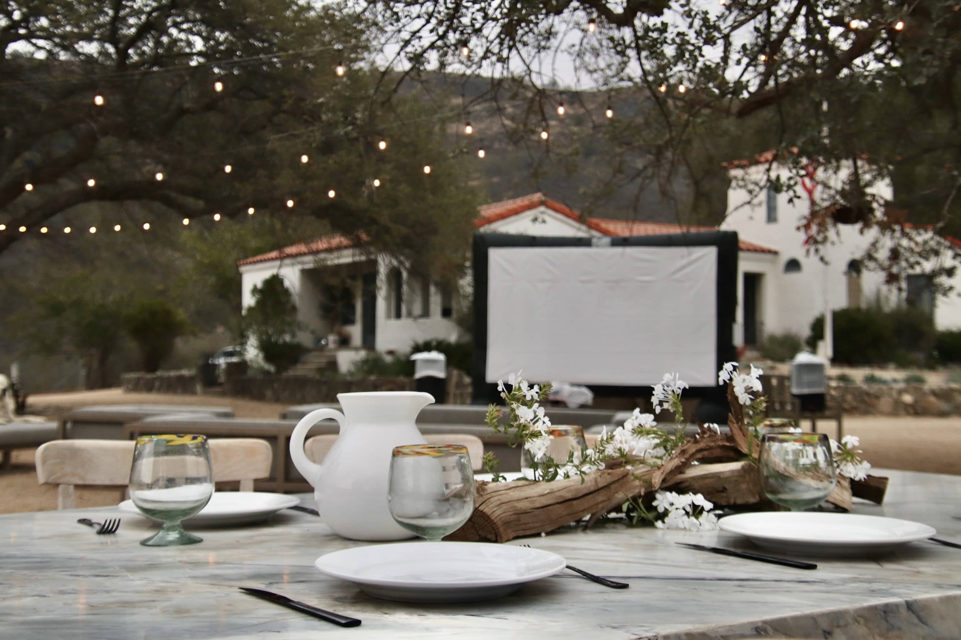 A rustic outdoor dining setup with a table adorned with plates, glasses, and a central arrangement of flowers, set against a backdrop of white buildings and string lights.