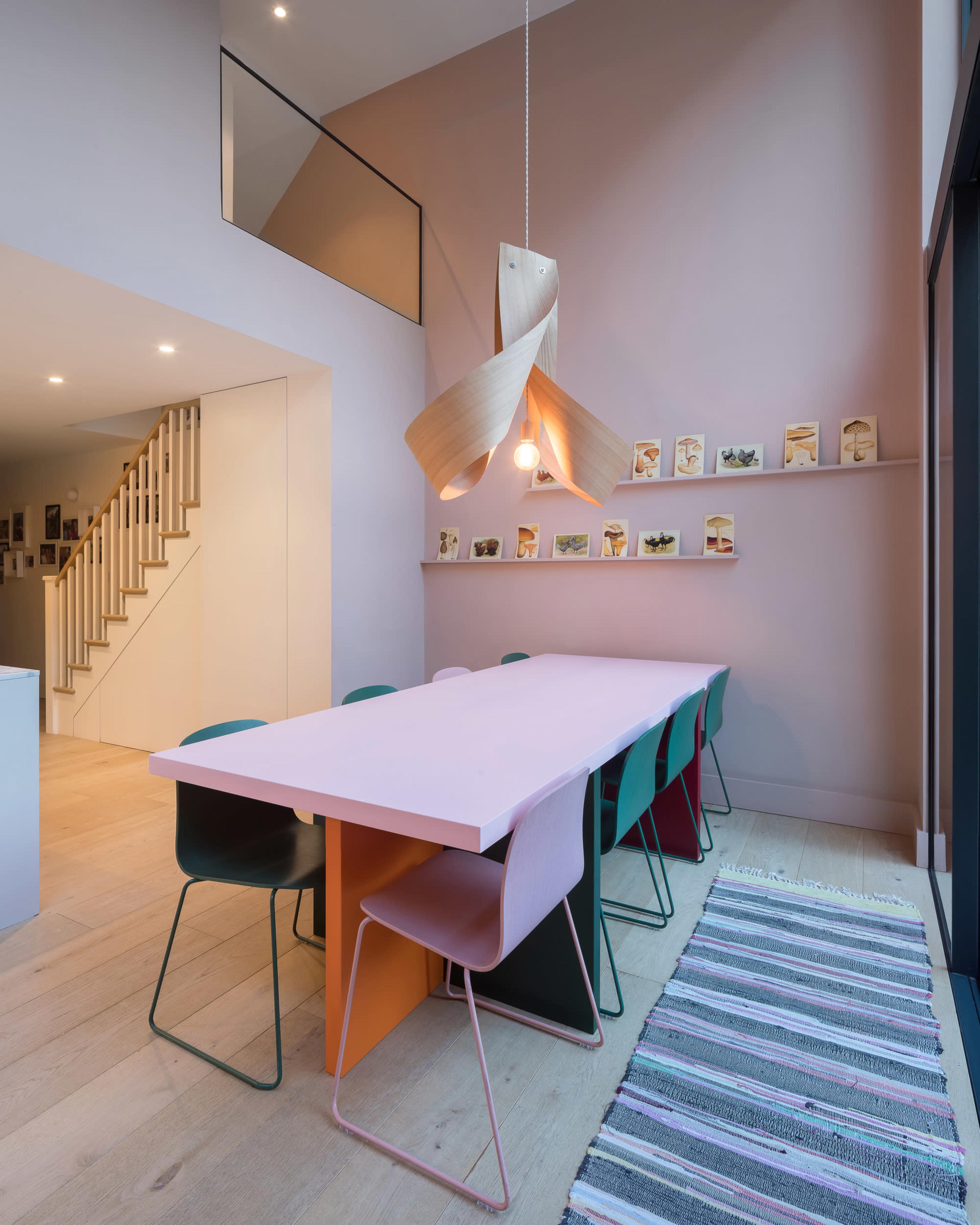 A dining area features a long pink table surrounded by multi-colored chairs and a wooden pendant light, with a gallery of framed artwork displayed on the wall.
