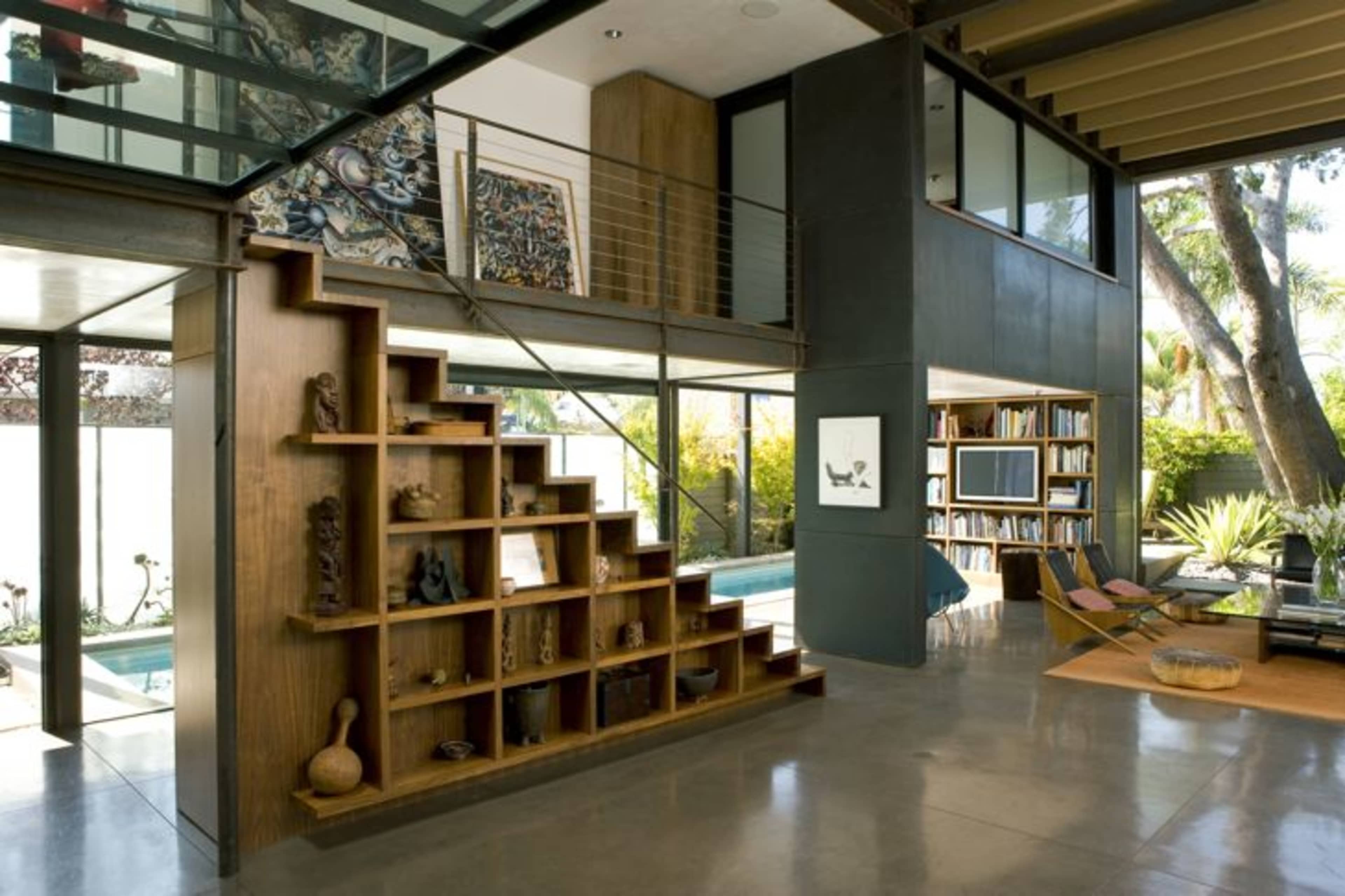 A modern interior featuring a staircase with built-in shelving, leading to an upper level overlooking a pool area surrounded by greenery.