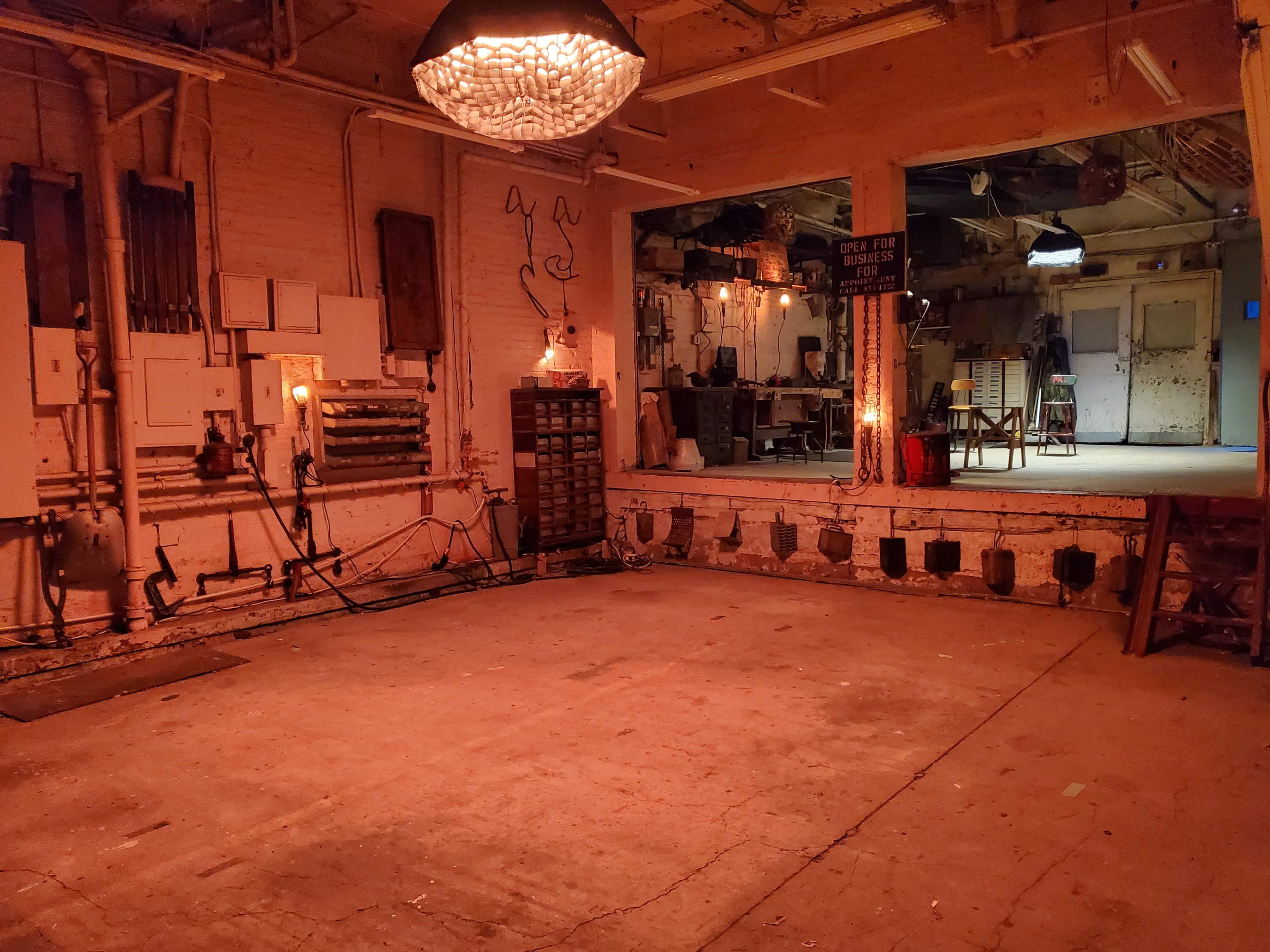 An industrial room with exposed pipes, a concrete floor, and a large light fixture hanging from the ceiling, leading to another room visible through an open doorway.