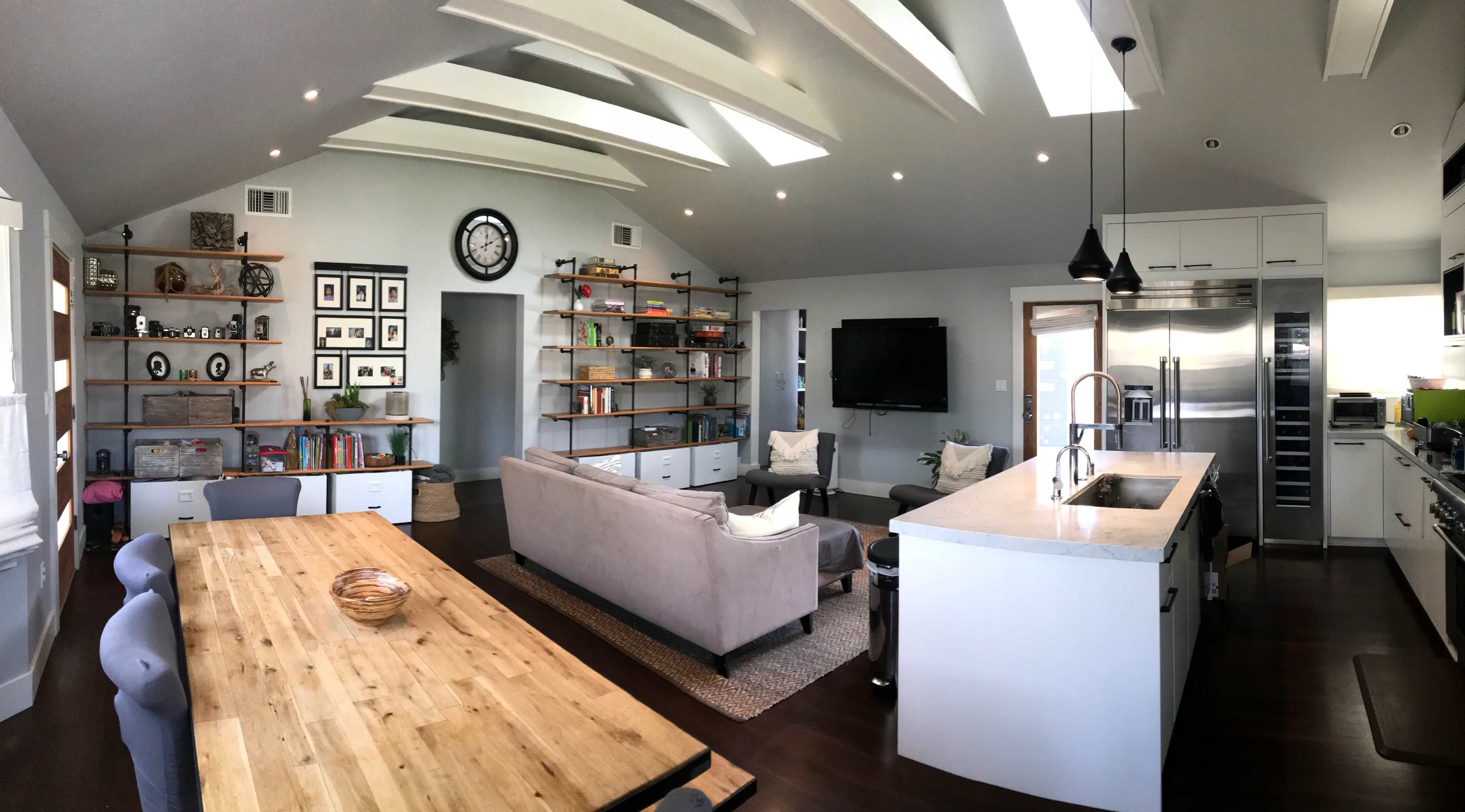 A modern open-concept living area with a kitchen, dining space, and a lounge that features shelves, a clock, and large windows.
