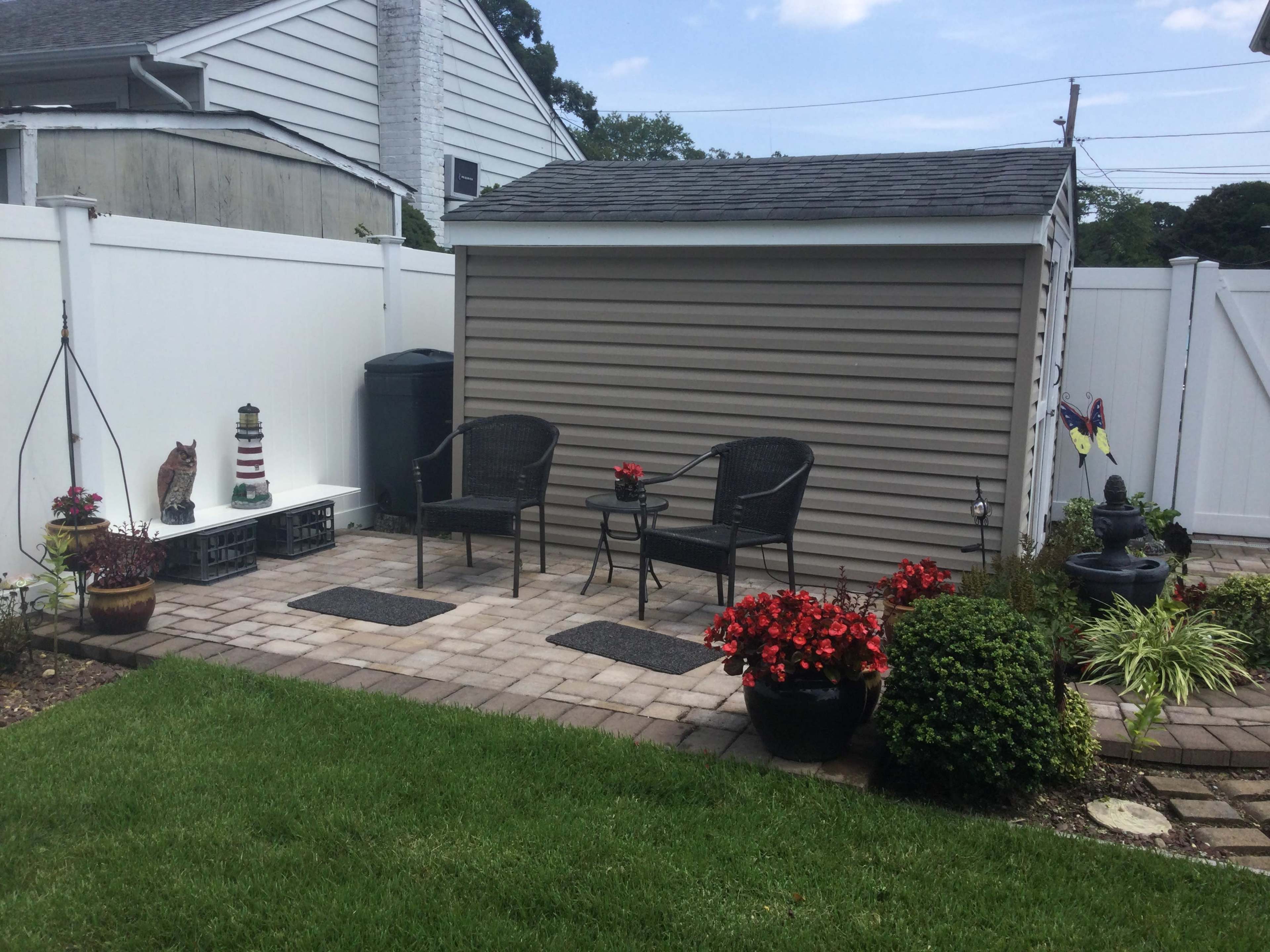 A backyard patio area with two black chairs, a small table, a storage shed, and various potted plants, all surrounded by a white fence.