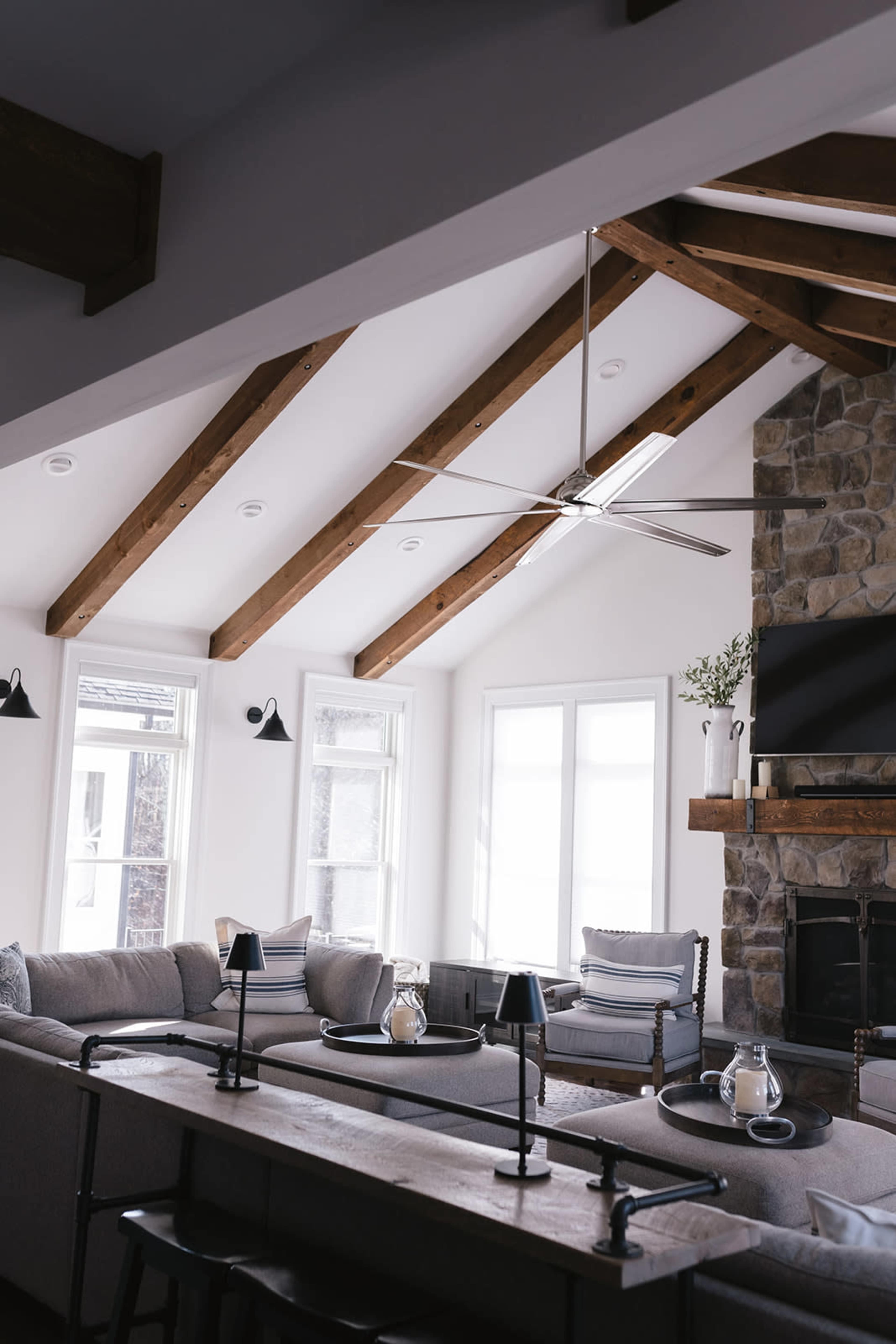 The living room features a stone fireplace, large windows, and exposed wooden beams, with a sectional sofa and coffee table in the center.