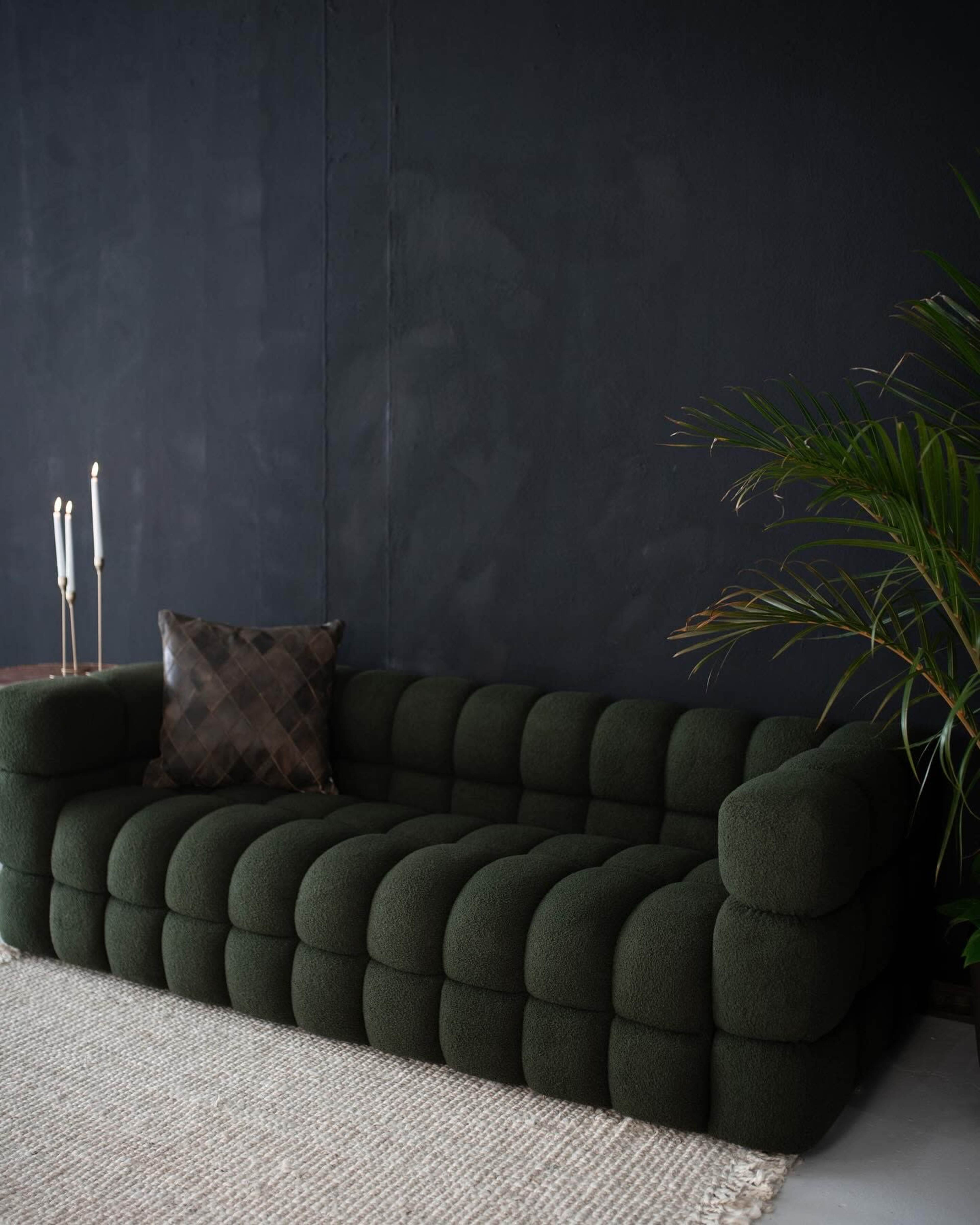 A green, tufted sofa is positioned against a dark wall, accompanied by a decorative pillow and a potted plant nearby.