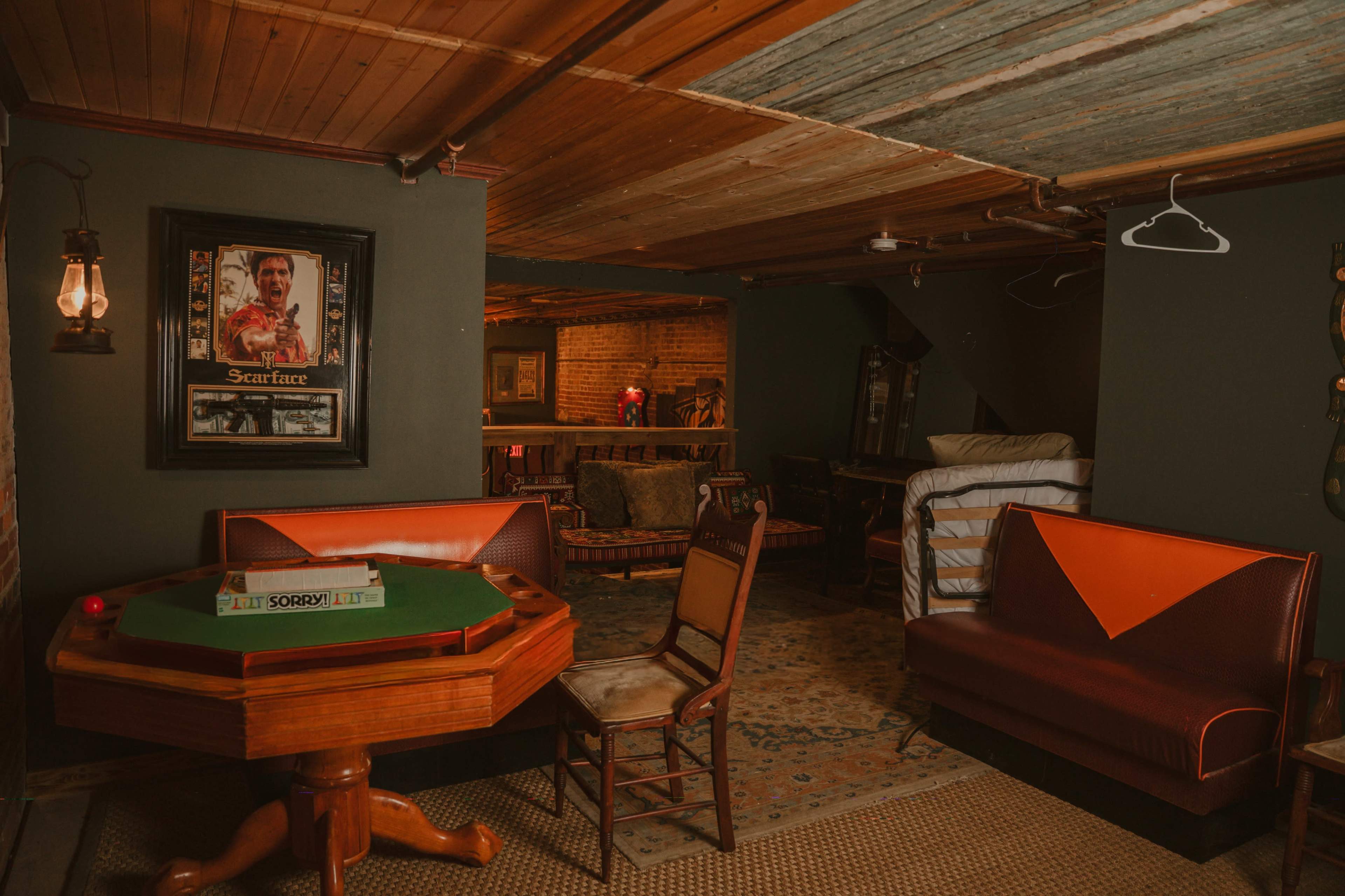 The image shows a dimly lit basement game room featuring a card table, wooden chairs, a vintage sofa, and a framed poster on the wall.
