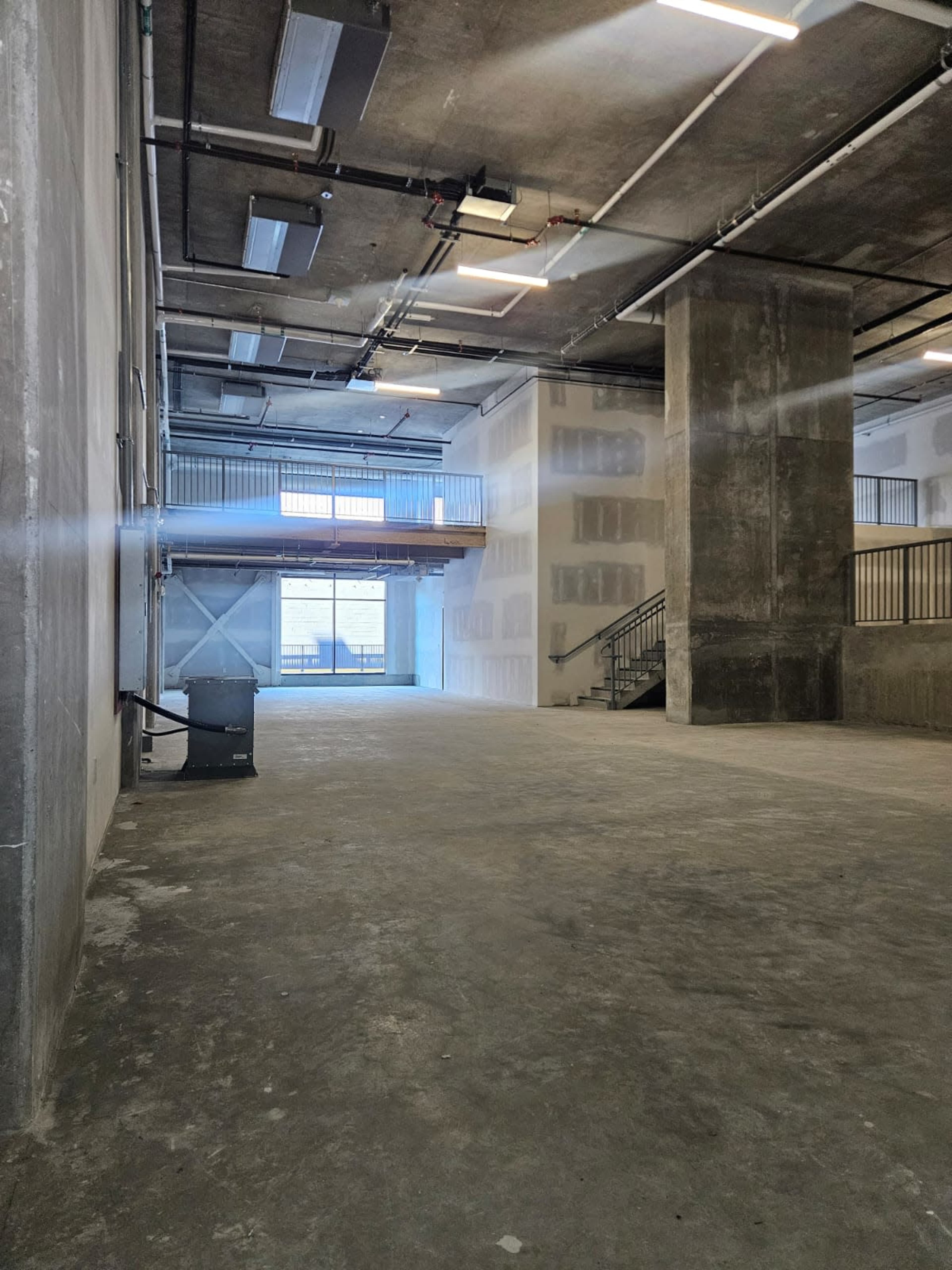 The image shows an empty, industrial-style interior space with concrete walls and floors, large windows allowing natural light, and a staircase in the background.