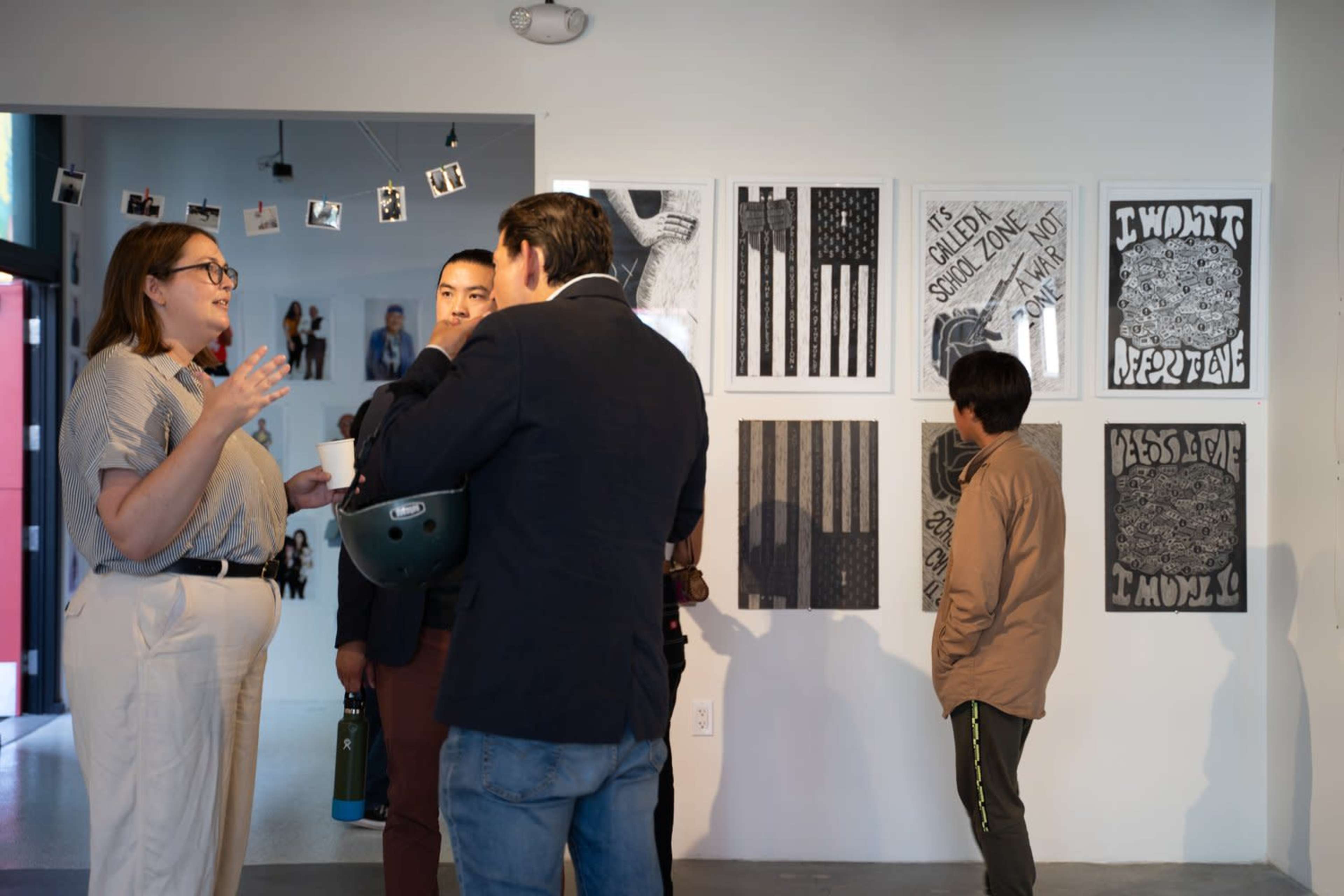 A group of people engage in conversation in an art gallery featuring several black-and-white artworks on the wall.