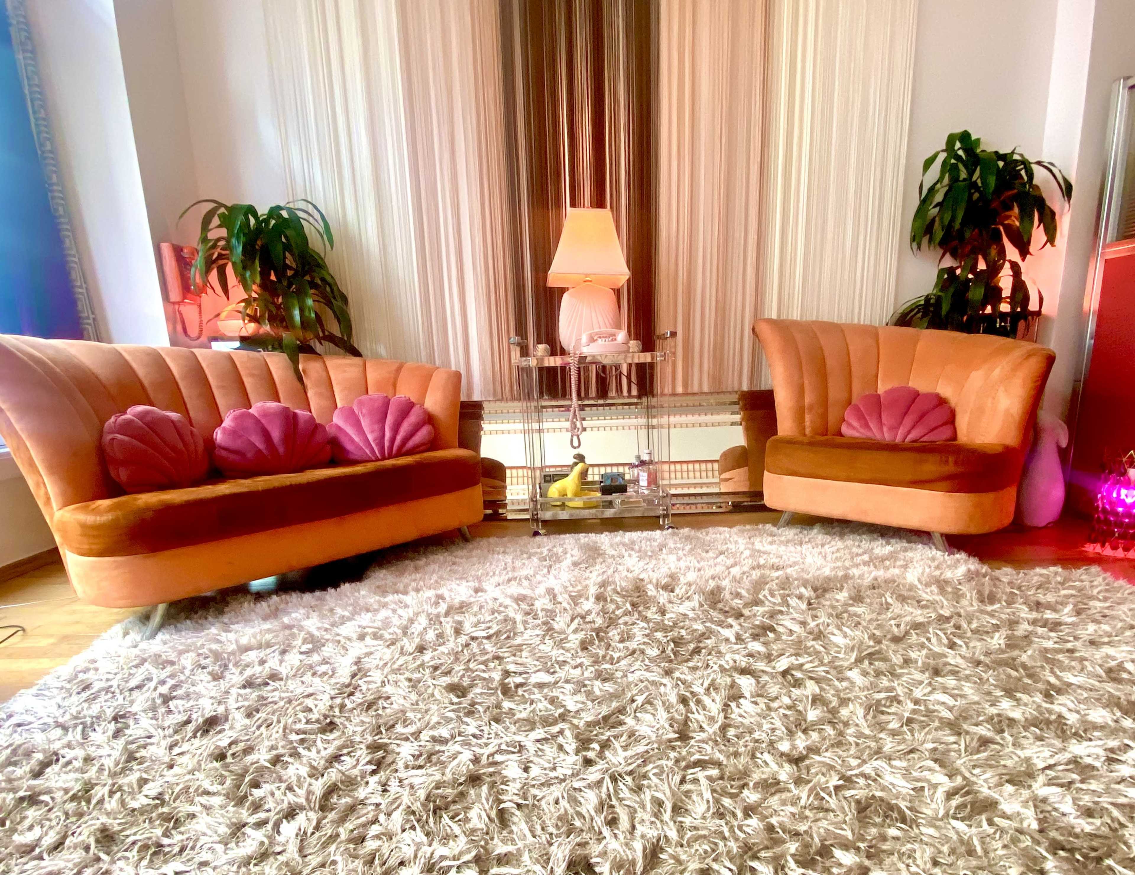 A living room with two orange velvet sofas, a glass coffee table, and a lamp, surrounded by plants and a shaggy white rug.