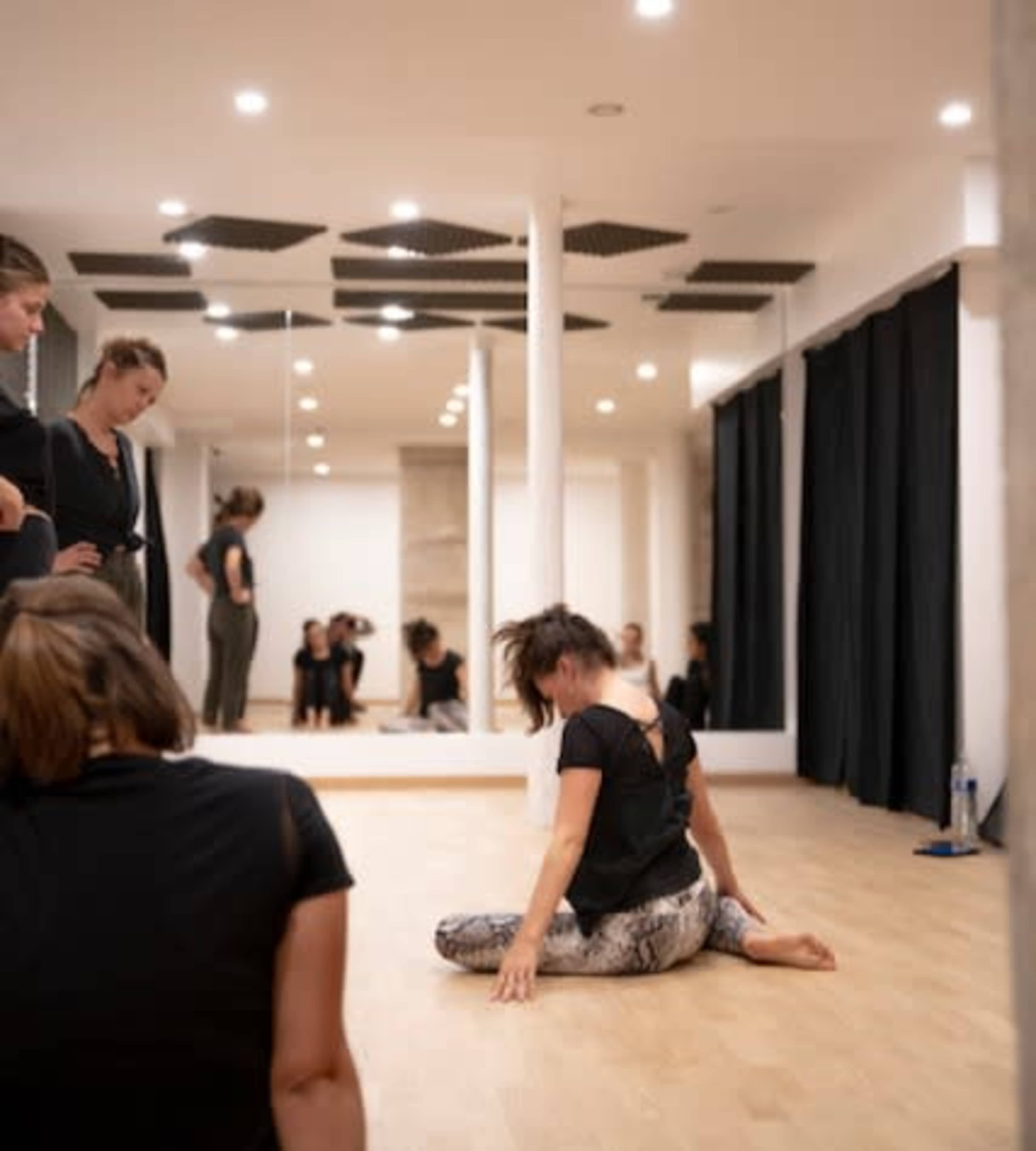 In a dance studio with mirrors, several individuals are observing one person performing a split on the floor.