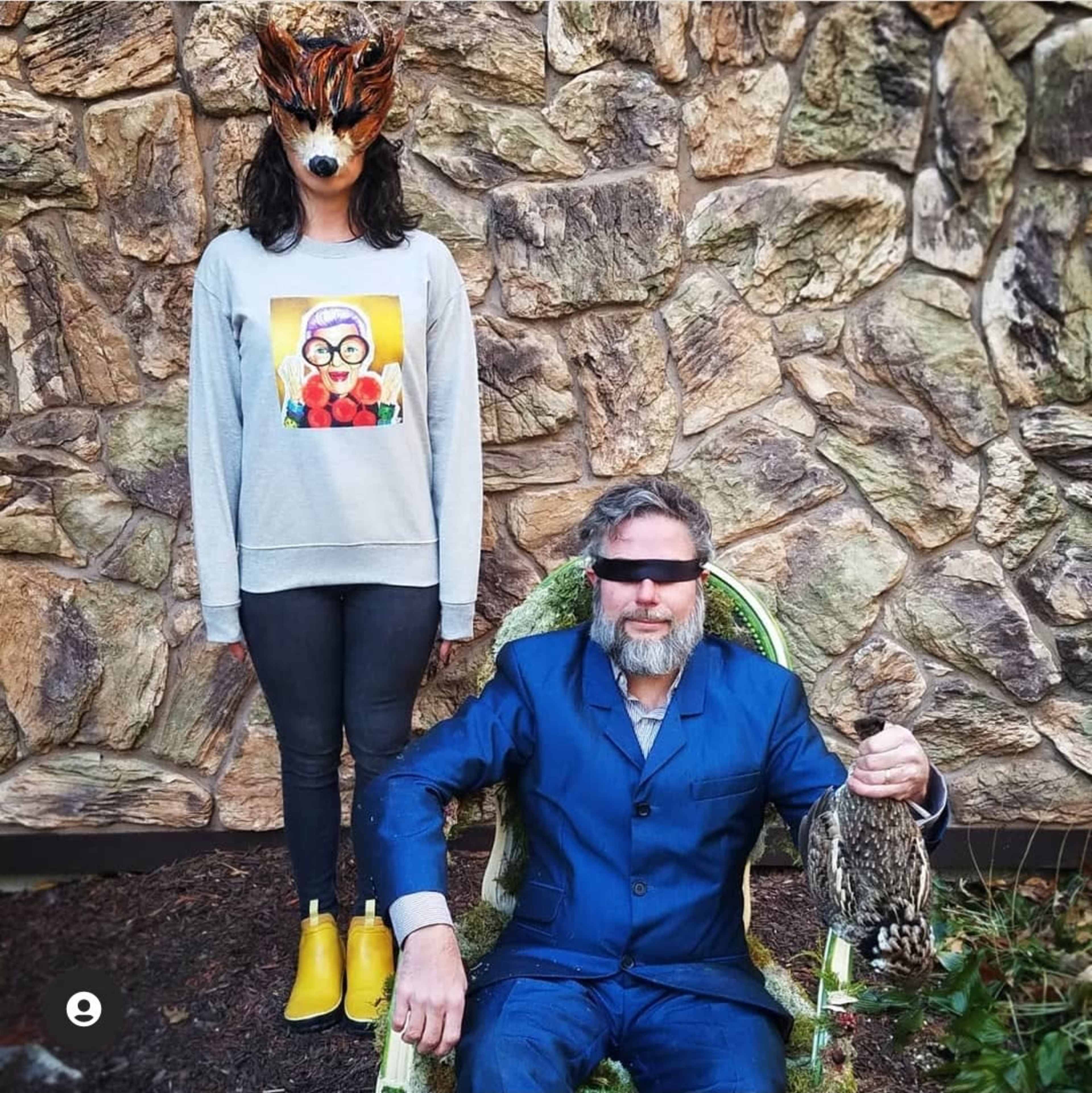 A woman wearing a fox mask stands behind a man in a blue suit, who is sitting in a chair and holding a bird, against a stone wall backdrop.