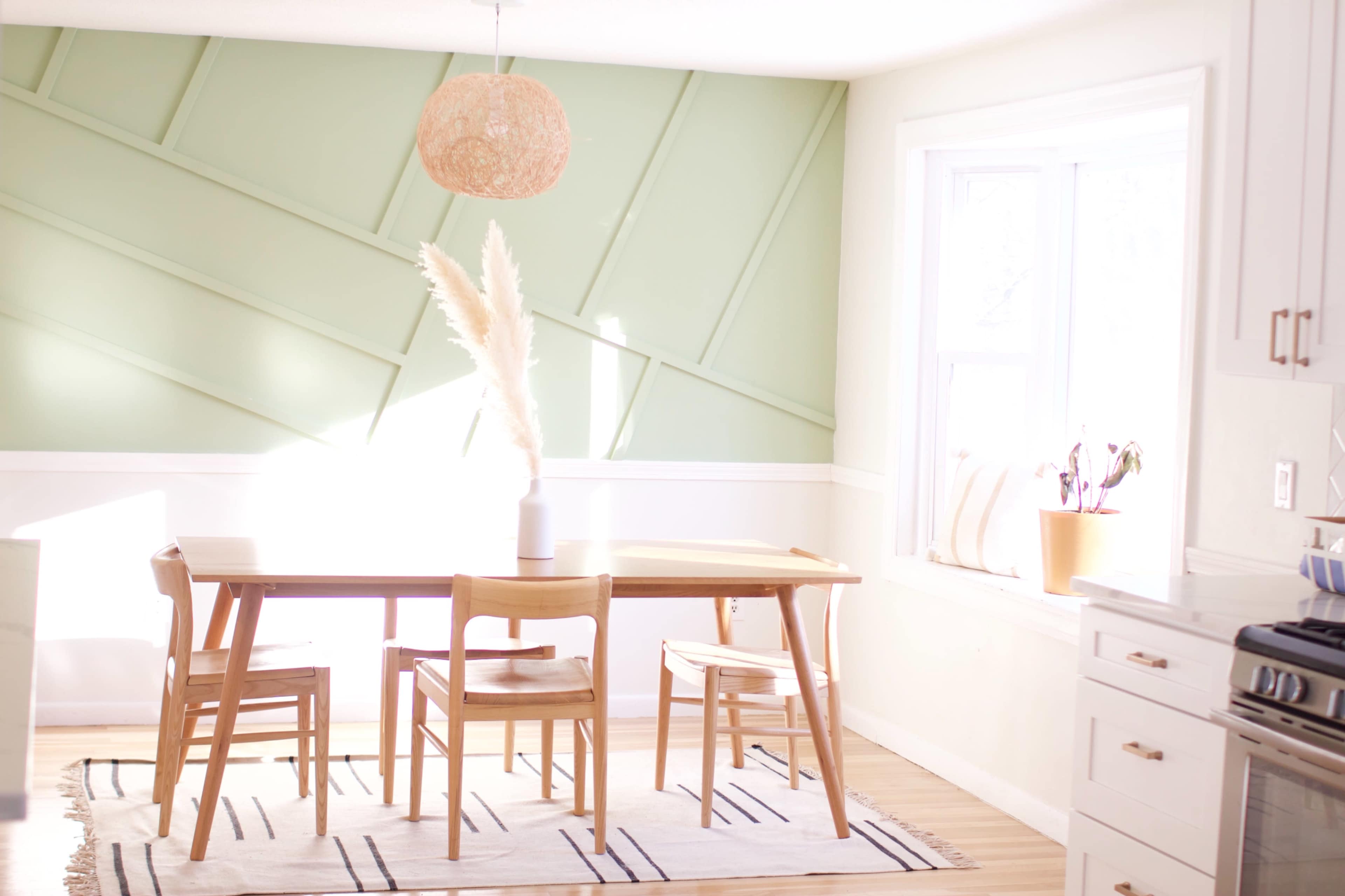 A wooden dining table with four chairs is positioned in a sunlit room featuring a green accent wall and a large window.