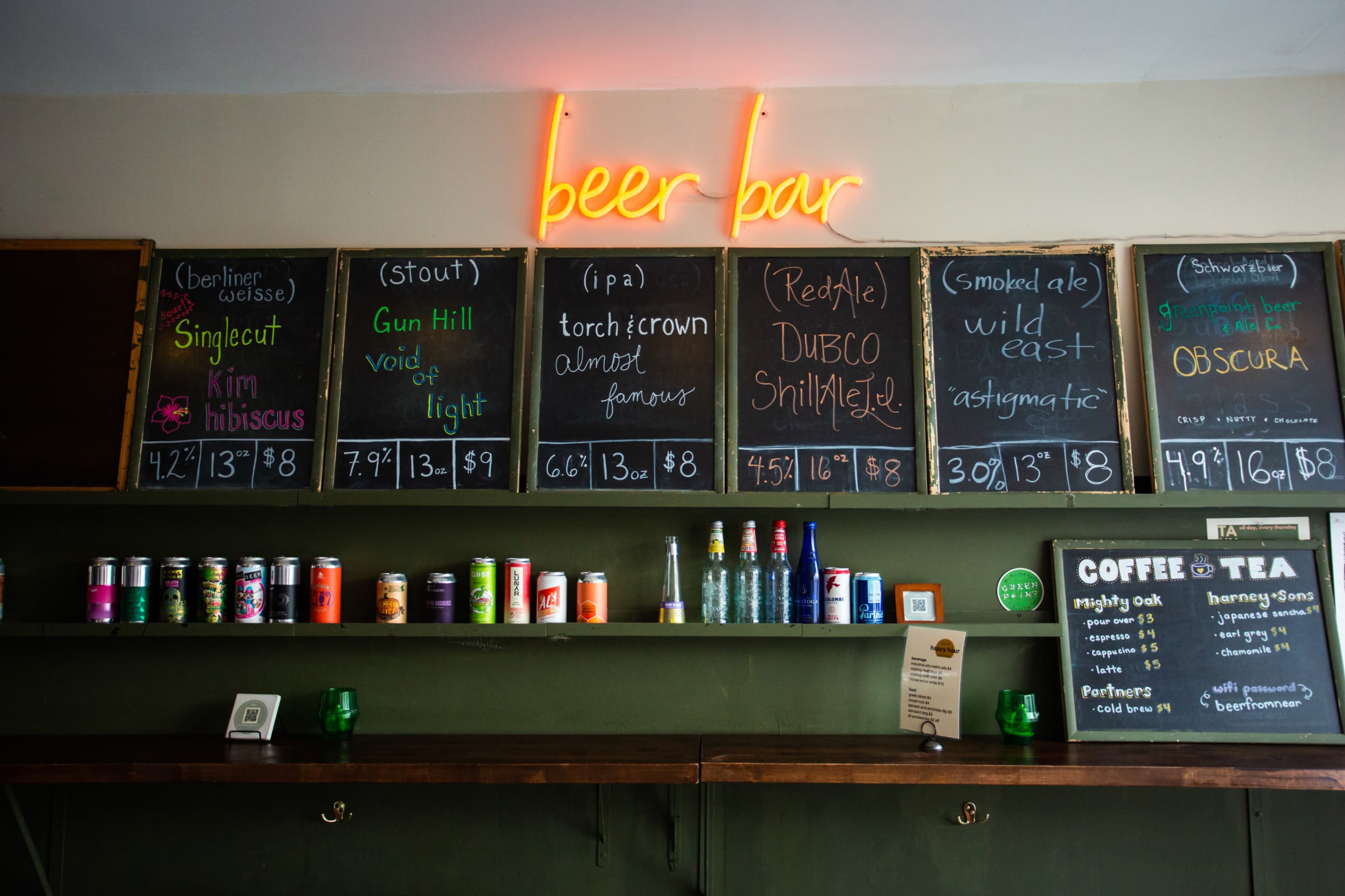 A beer bar with a neon sign that reads "beer bar" and a chalkboard menu listing various beers and their prices, alongside cans and bottles displayed on shelves.