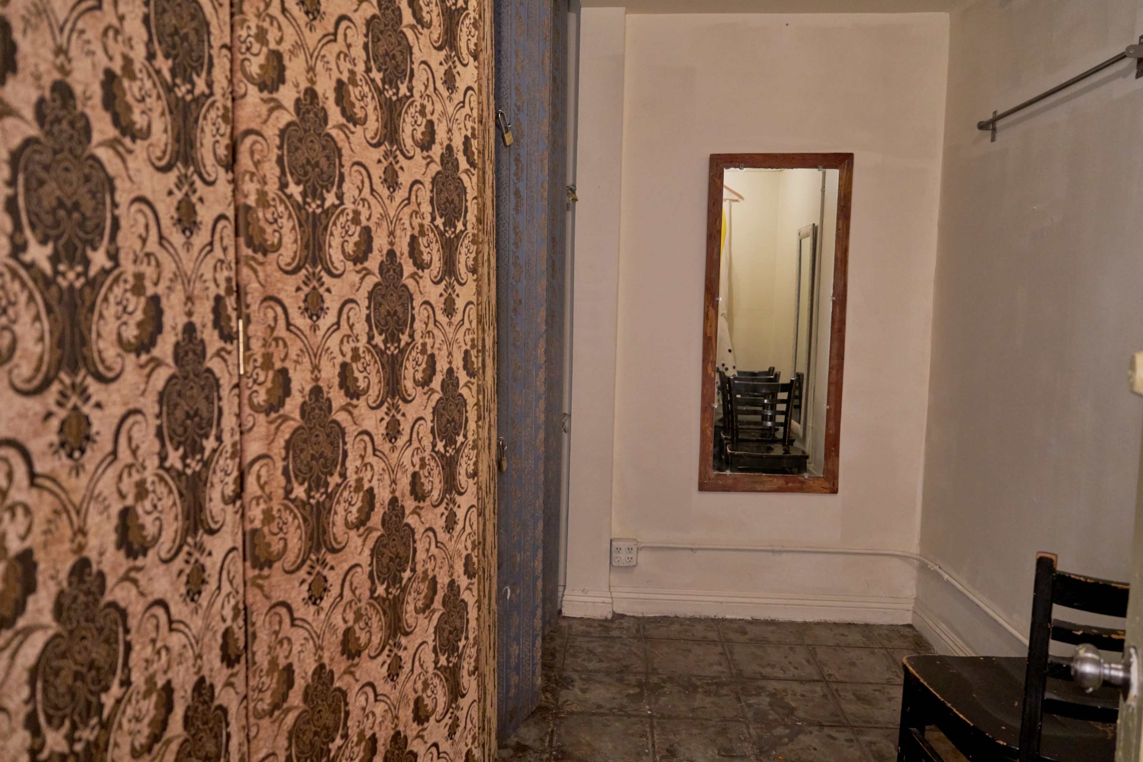 The image shows a narrow, empty room with patterned wallpaper on one side, a mirror on another wall, and plain tiled flooring.