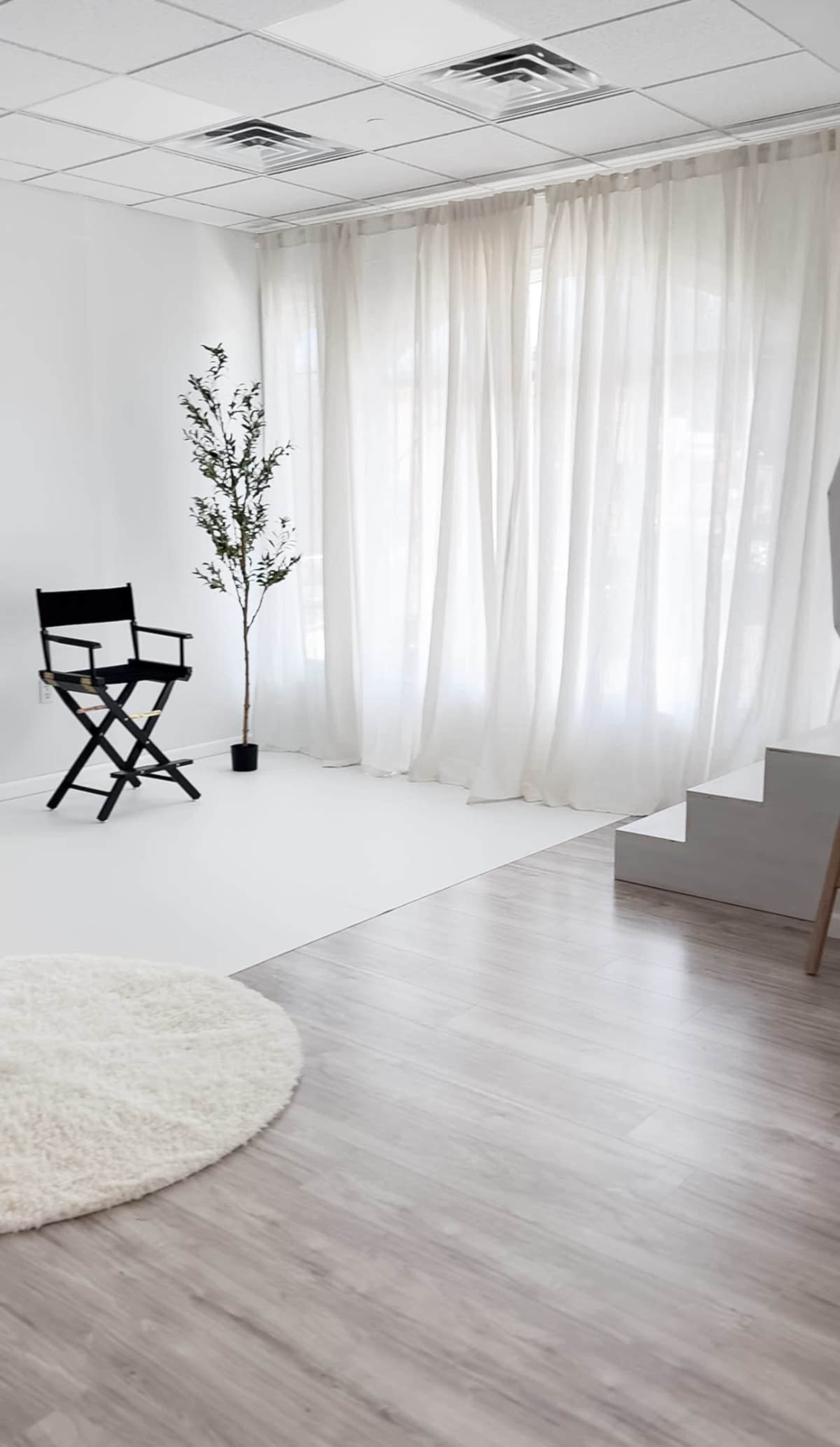 The image shows a bright, minimalistic room featuring a black director's chair, a small plant in a pot, and large white curtains beside a staircase.