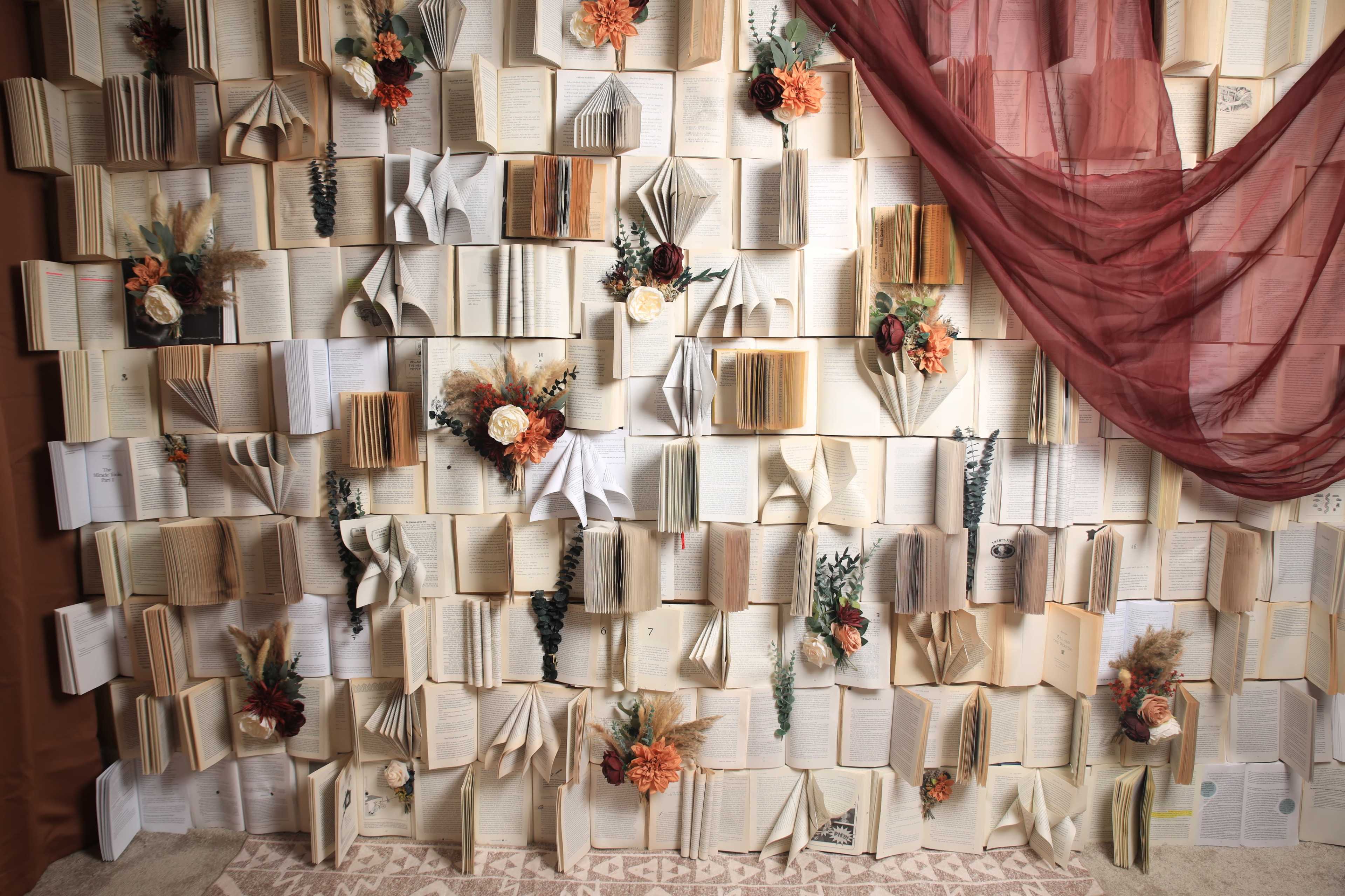 The image shows a decorative wall made of open books arranged in various positions, complemented by fabric drapes and artificial flowers.