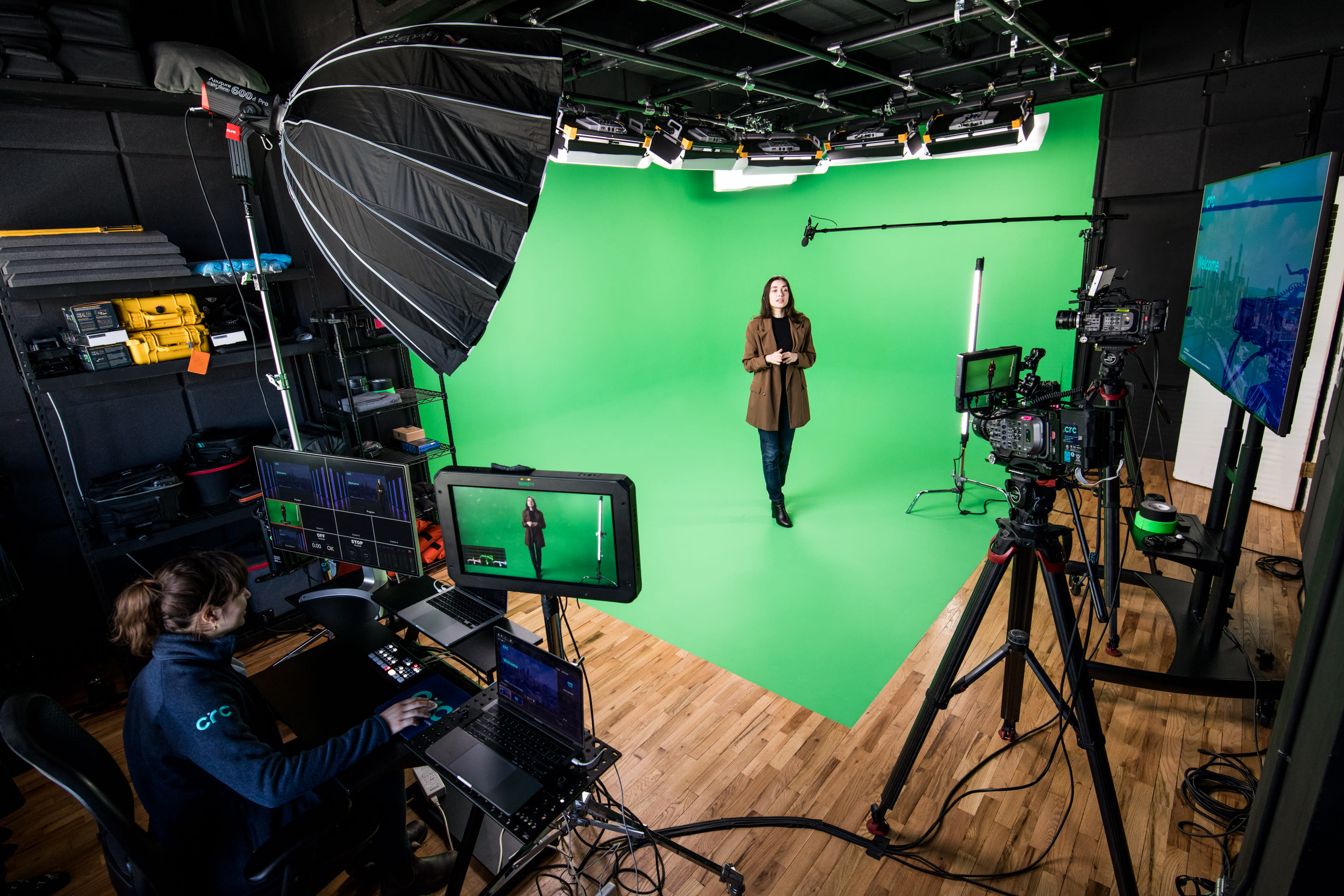 A woman stands in front of a green screen while a person operates equipment at a nearby workstation filled with monitors and cameras.