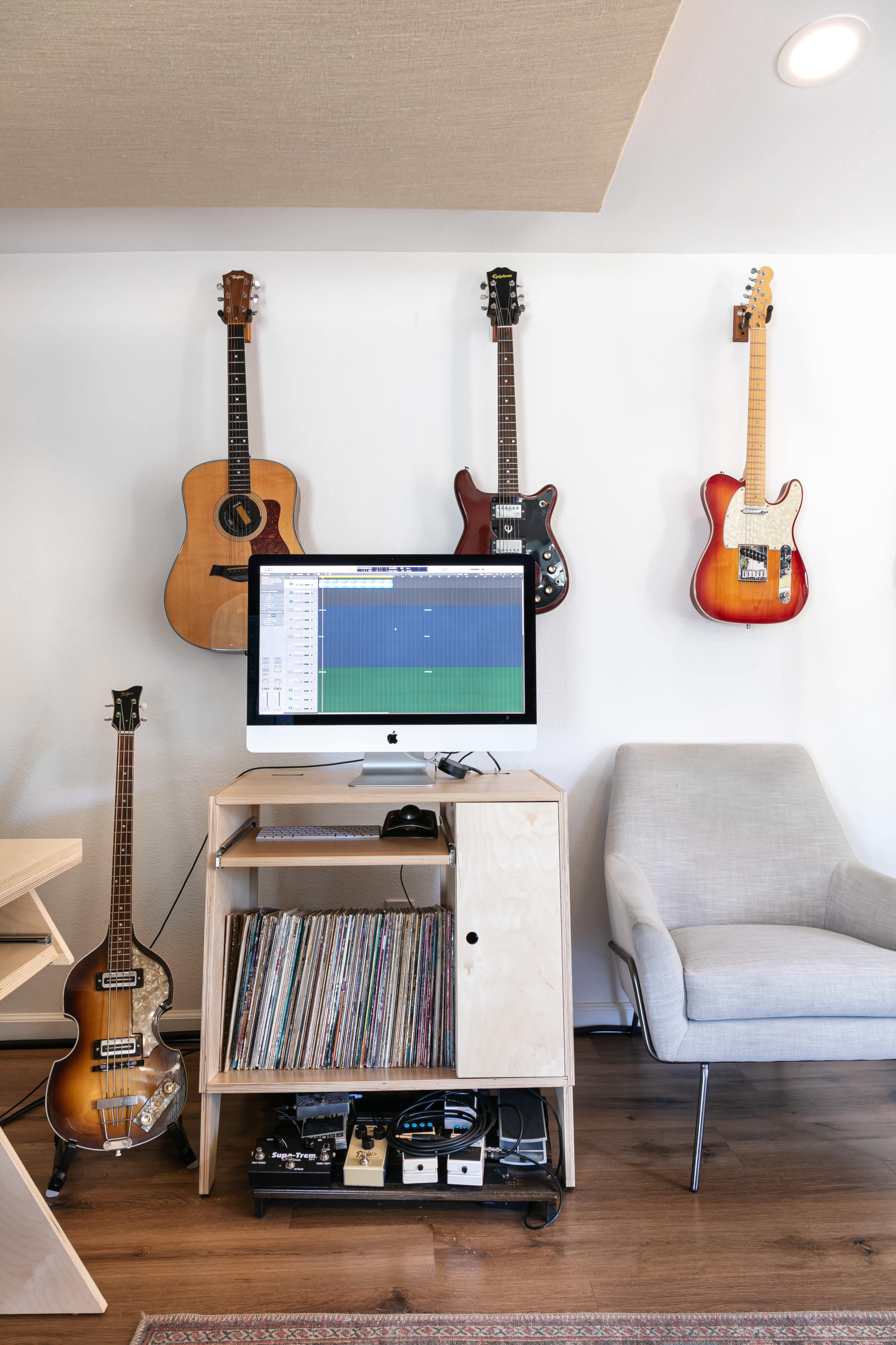 A workspace features a computer with music software, surrounded by various guitars on the wall and a cabinet holding vinyl records and music equipment.