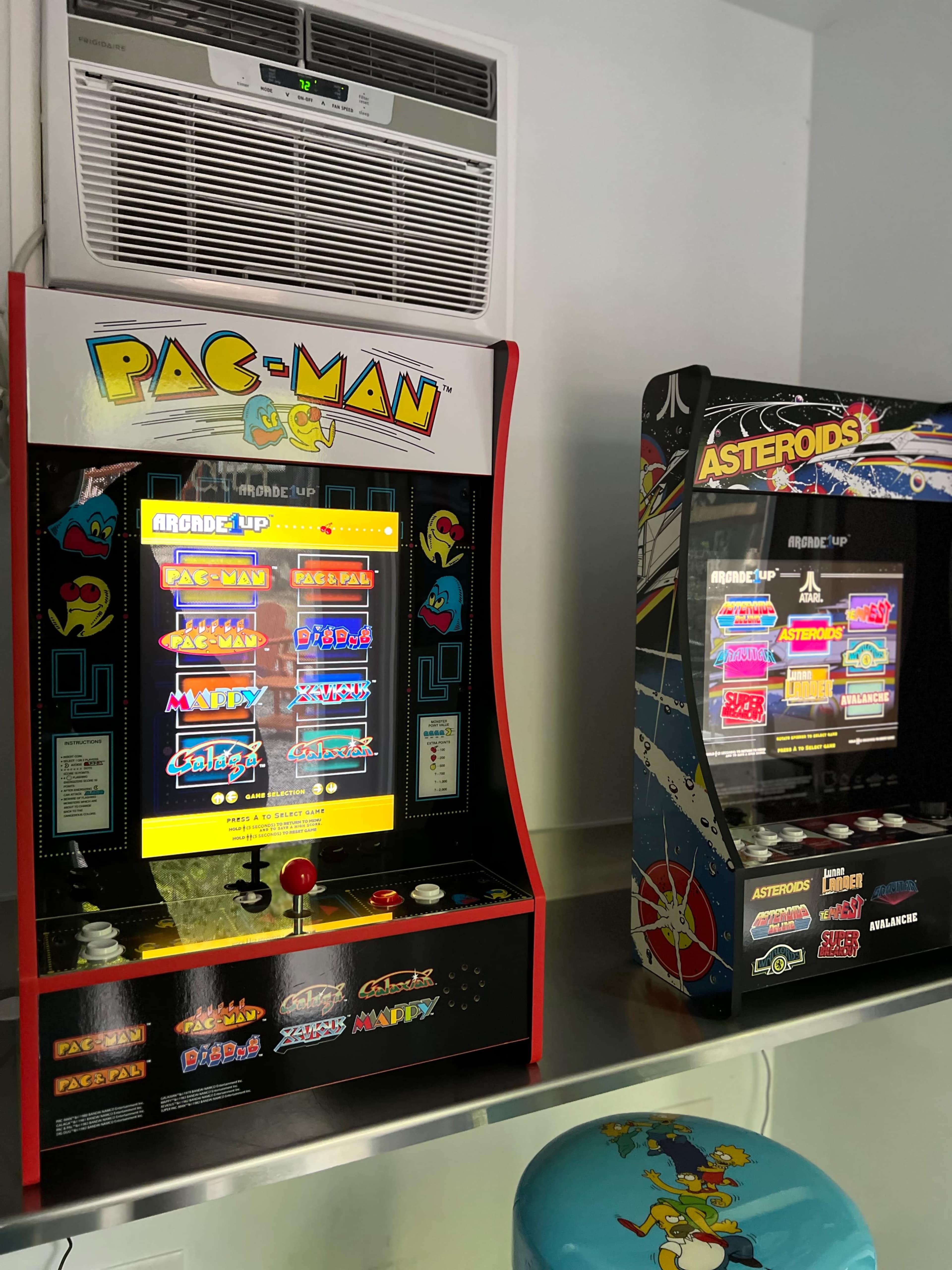 Two arcade game machines, one featuring Pac-Man and the other Asteroids, placed side by side in a brightly lit room.