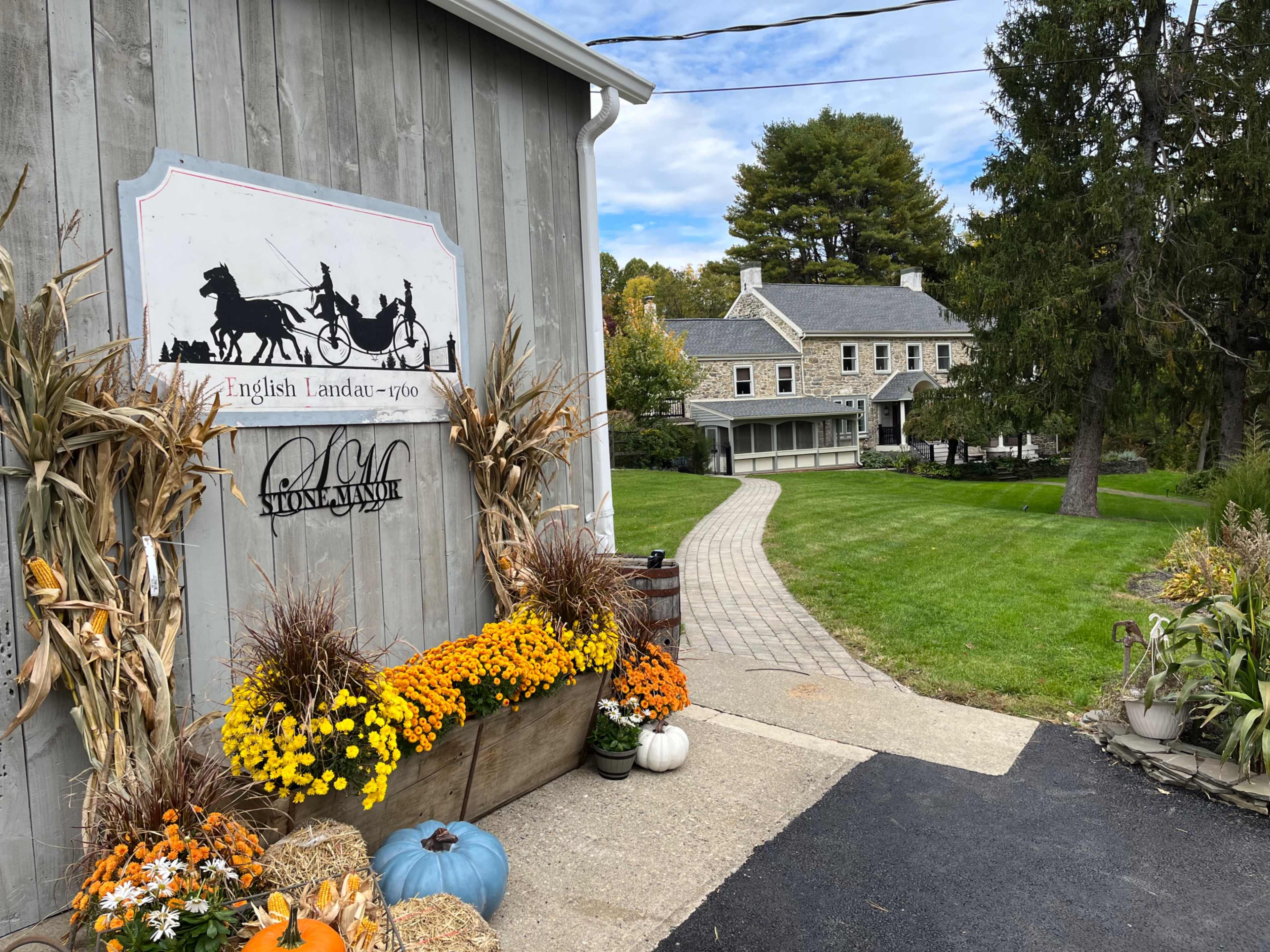 A stone house with a gray exterior is surrounded by landscaped grounds, featuring a curved path and decorative autumn plants near the entrance.