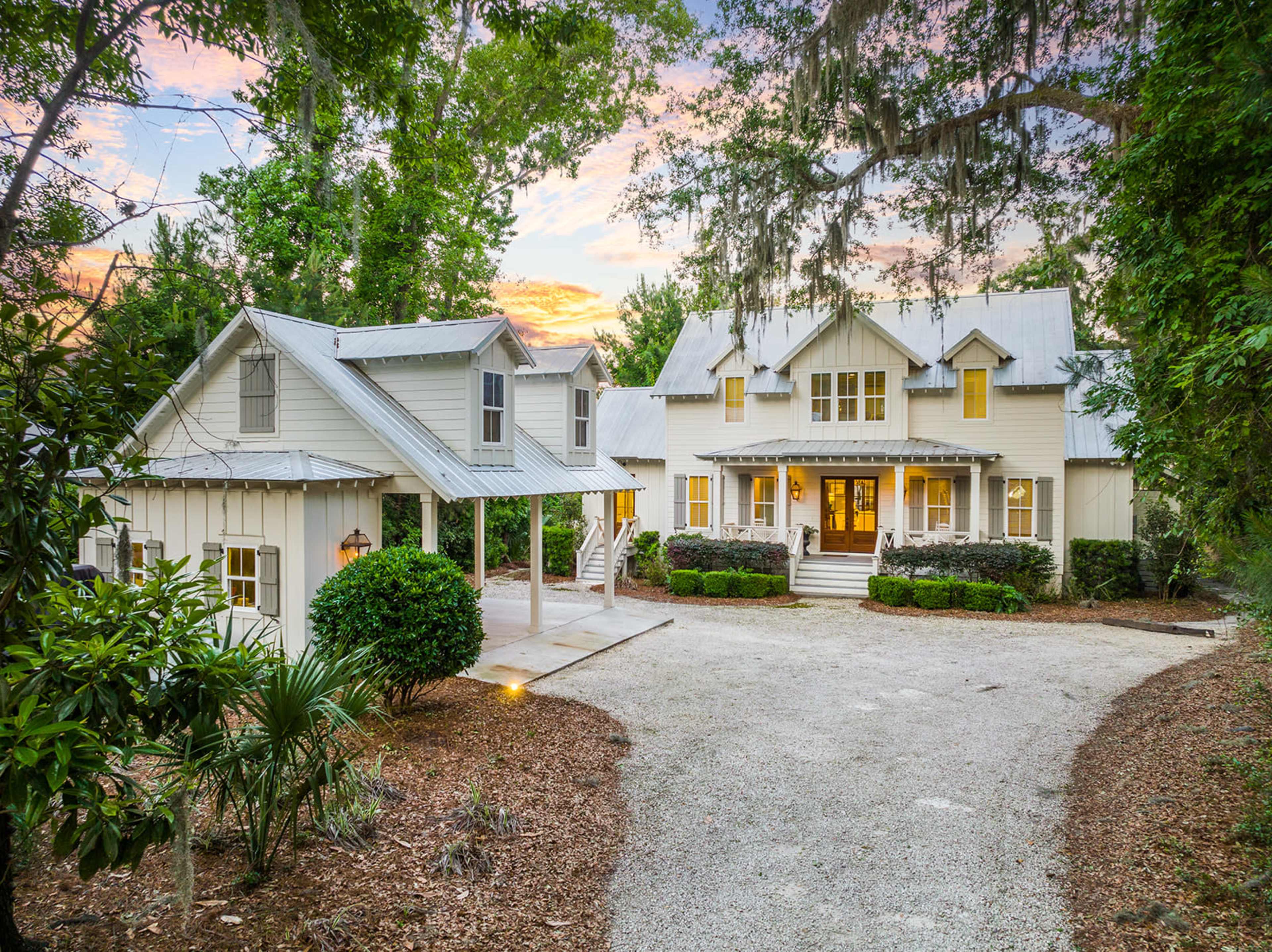 A white, two-story house with a front porch and multiple gabled roofs is set among trees and landscaped greenery in a gravel driveway.