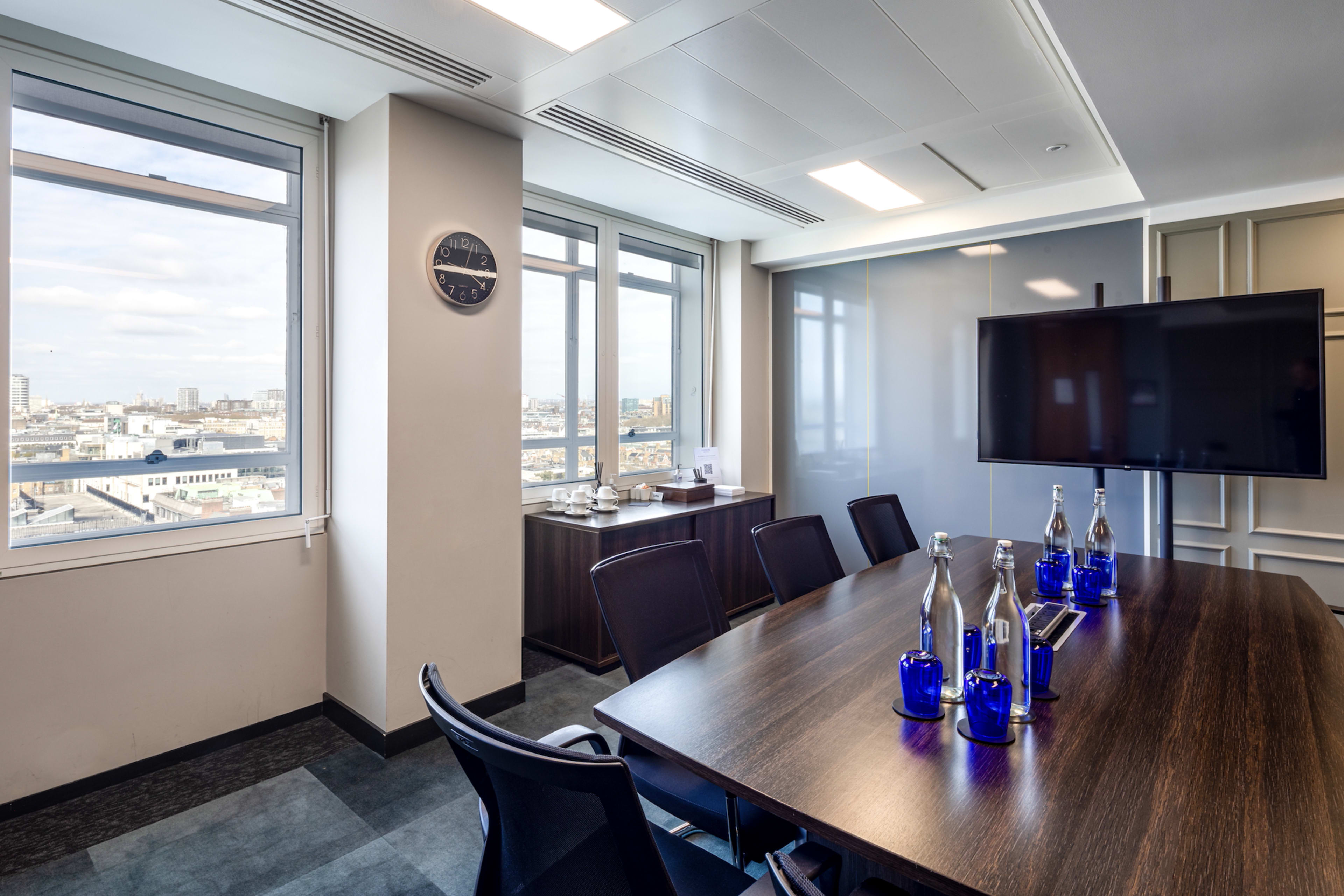 A modern conference room features a large wooden table, several glass water bottles, a wall-mounted television, and views of the city through large windows.