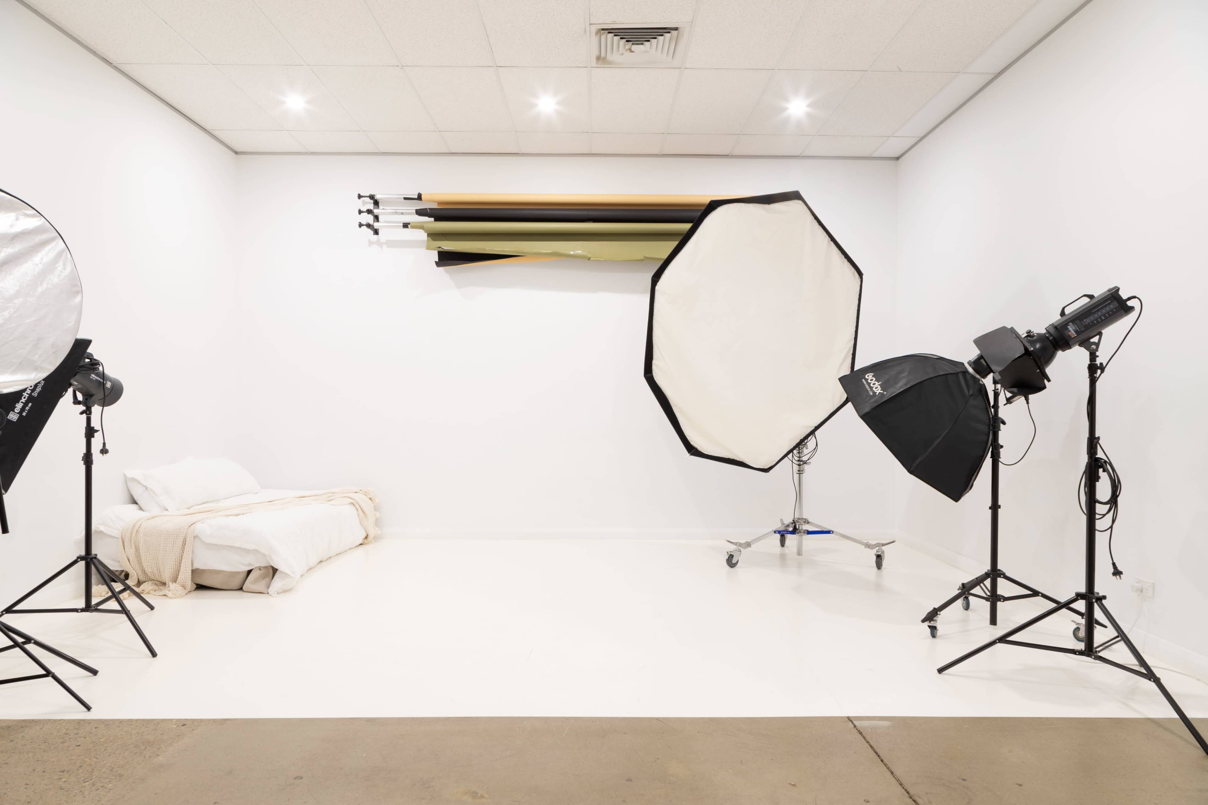 A photography studio features a bed with a light blanket, several backdrops mounted on the wall, and various lighting equipment arranged around the space.