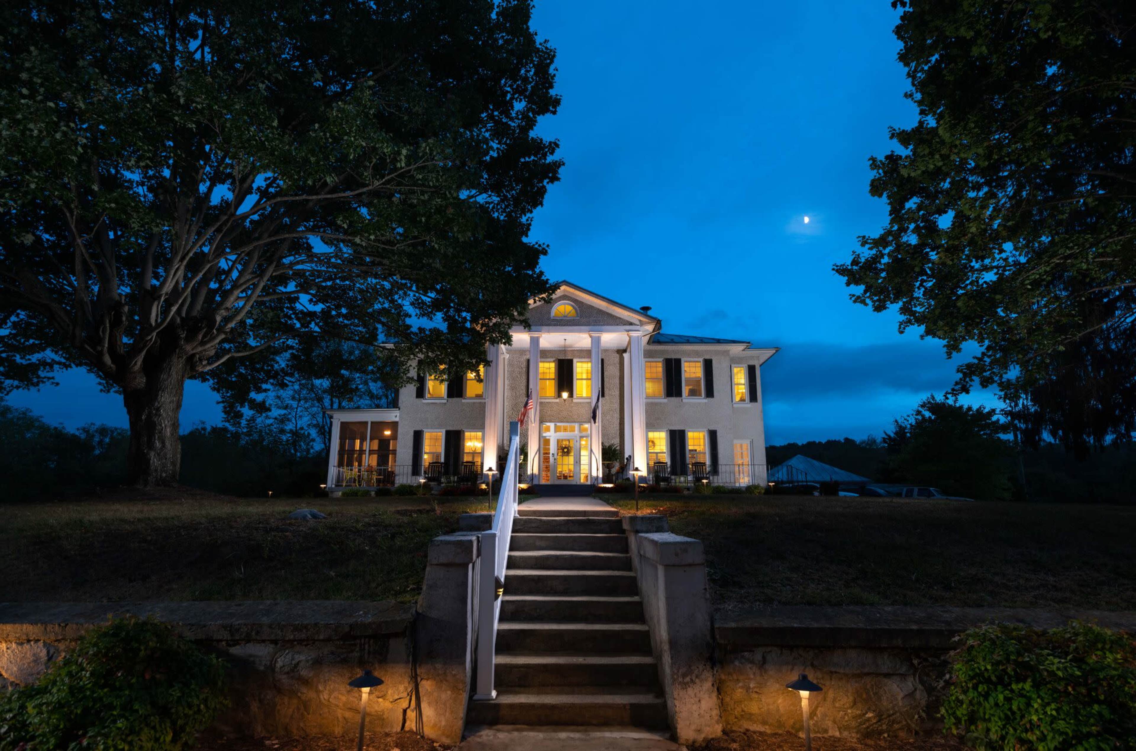 A large two-story house with white siding and bright windows stands at the top of a landscaped staircase, illuminated against a twilight sky.