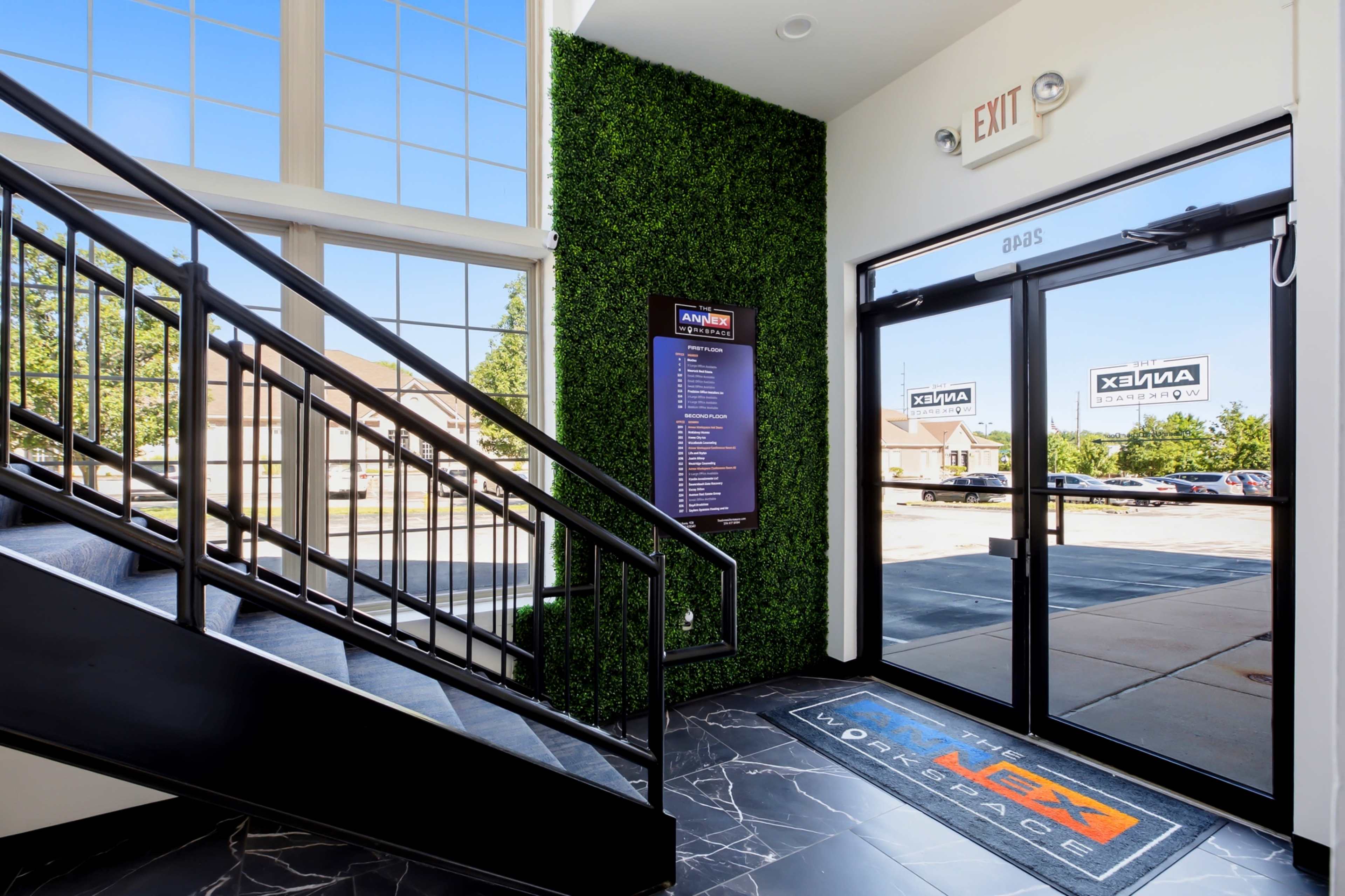 The image shows a modern entrance area featuring a glass door, a staircase with a black railing, and a green wall, along with a directory sign on display.