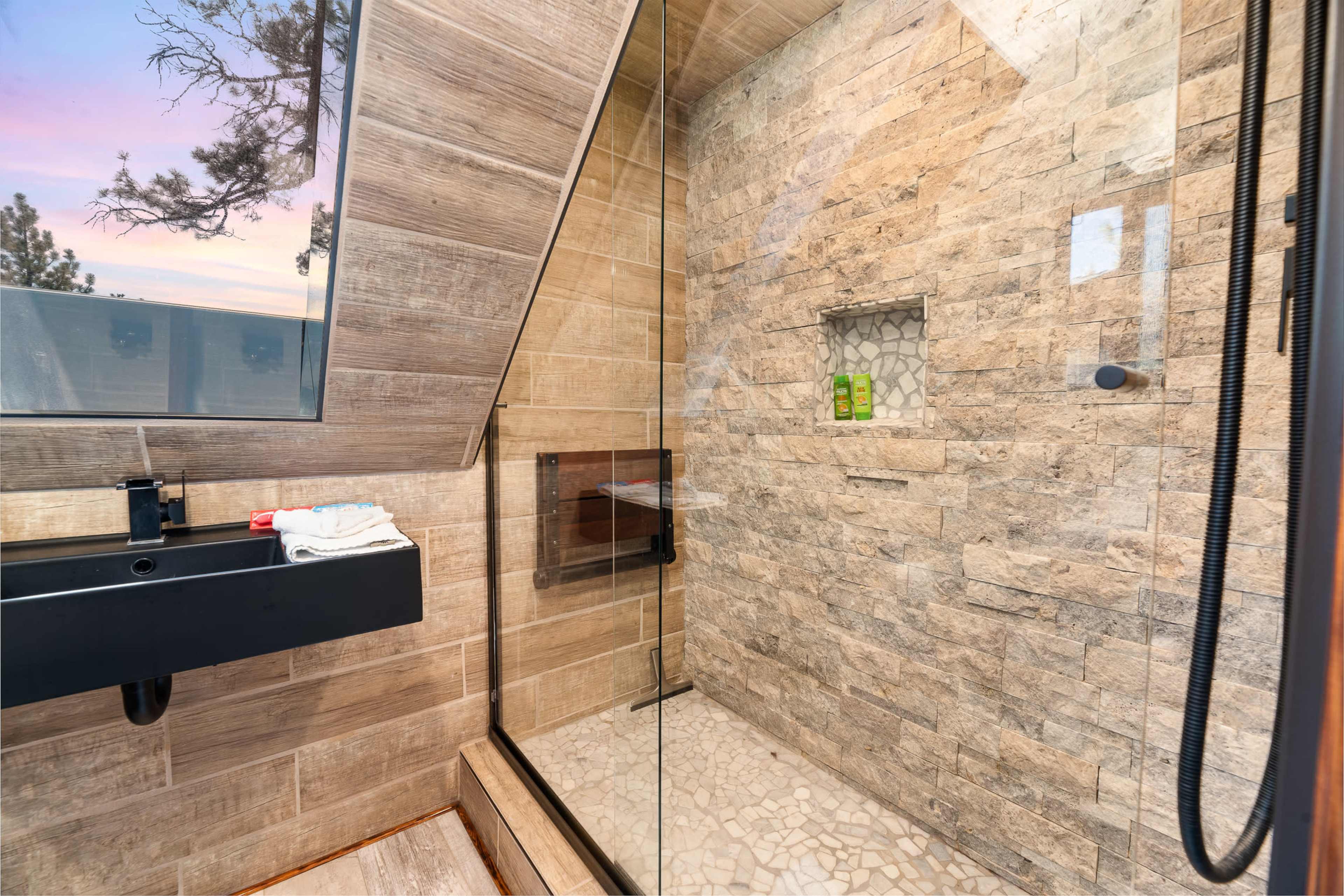 A modern shower enclosed with glass, featuring stone walls and a wooden sink.