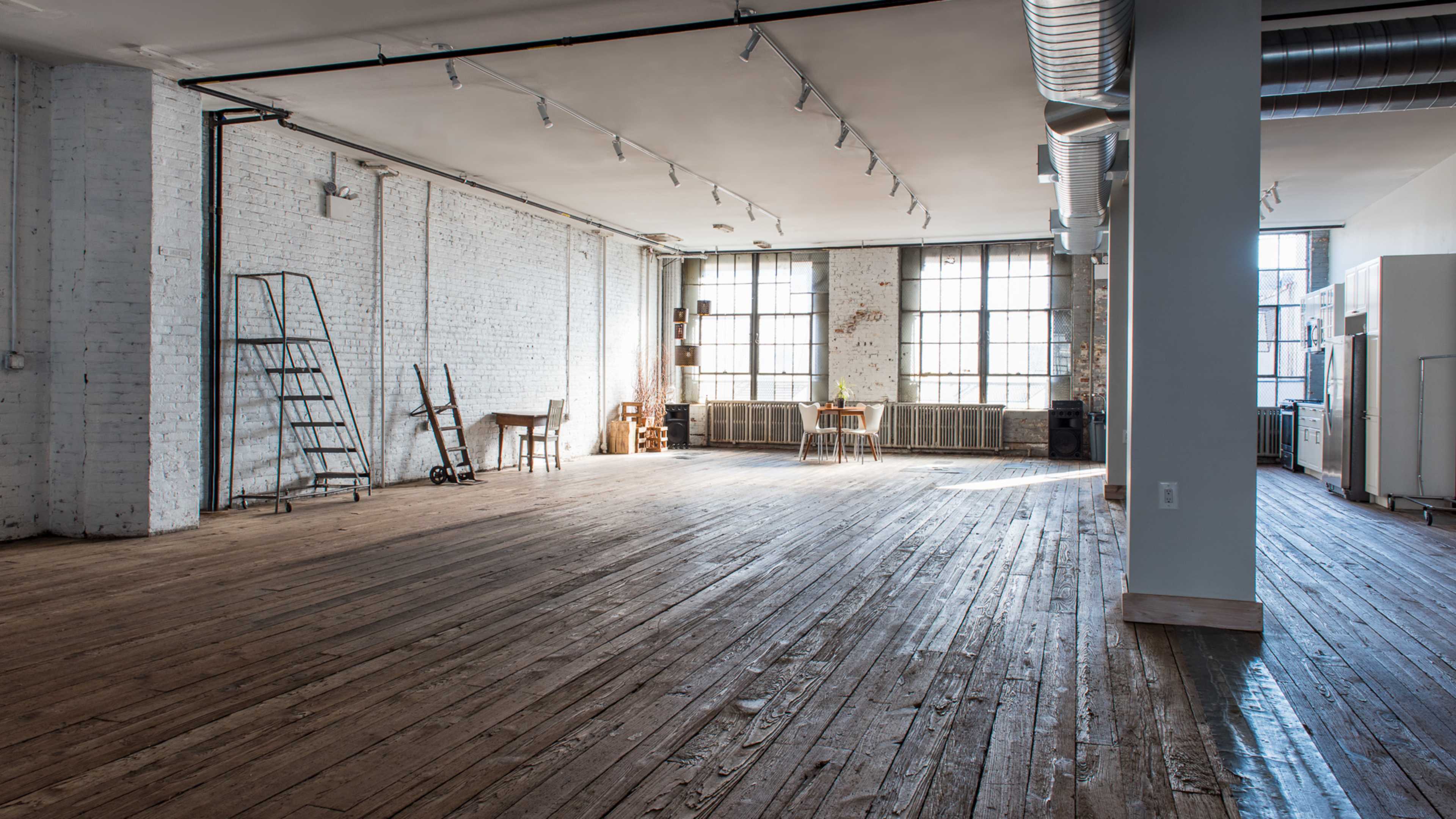 A spacious, empty room with large windows, exposed brick walls, and wooden flooring, featuring minimal furniture such as a table and a chair.