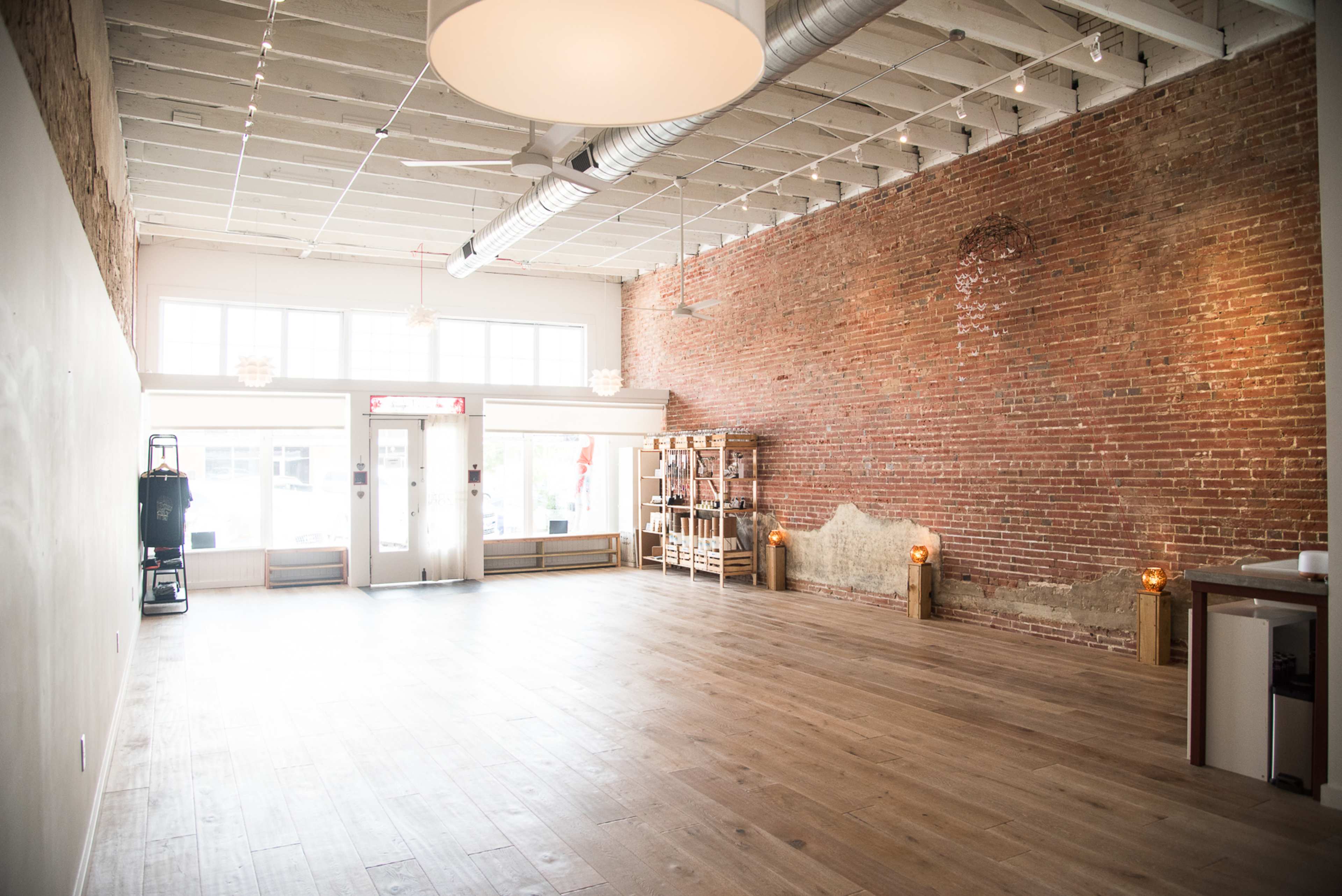 The image shows a spacious, vacant interior with exposed brick walls, wooden flooring, and large windows letting in natural light.